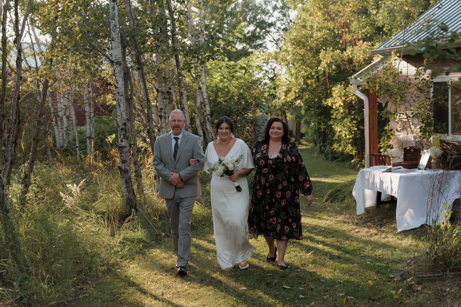 A tranquil outdoor wedding ceremony in a wooded setting with guests walking along a sun-dappled path.