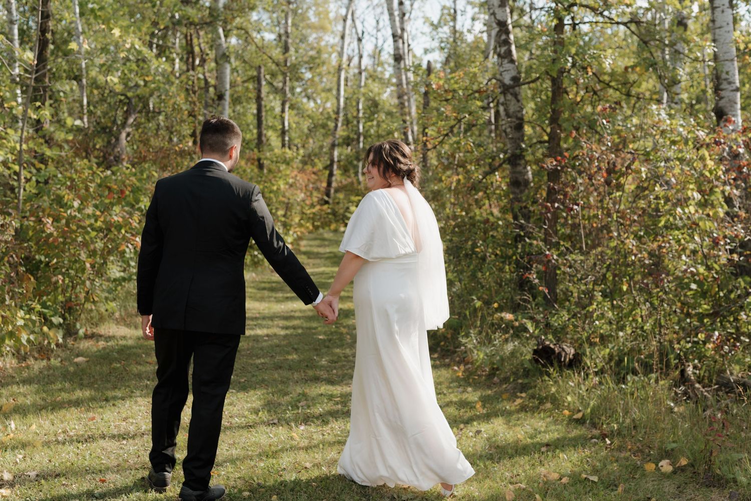 A couple in wedding attire walking hand in hand through a sunlit wooded area on their special day.
