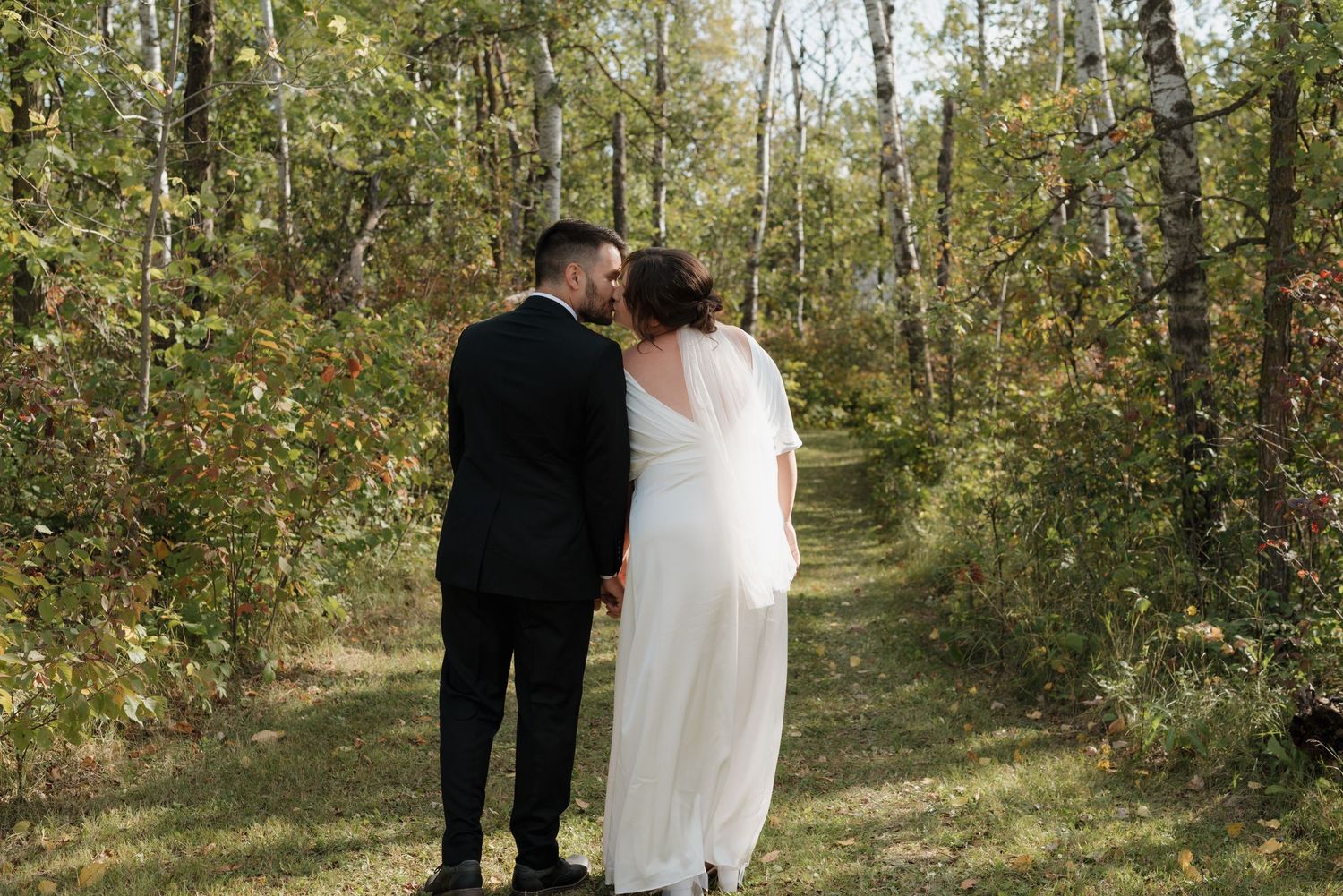 A couple in wedding attire stand together on a sunlit forest path surrounded by autumn trees.