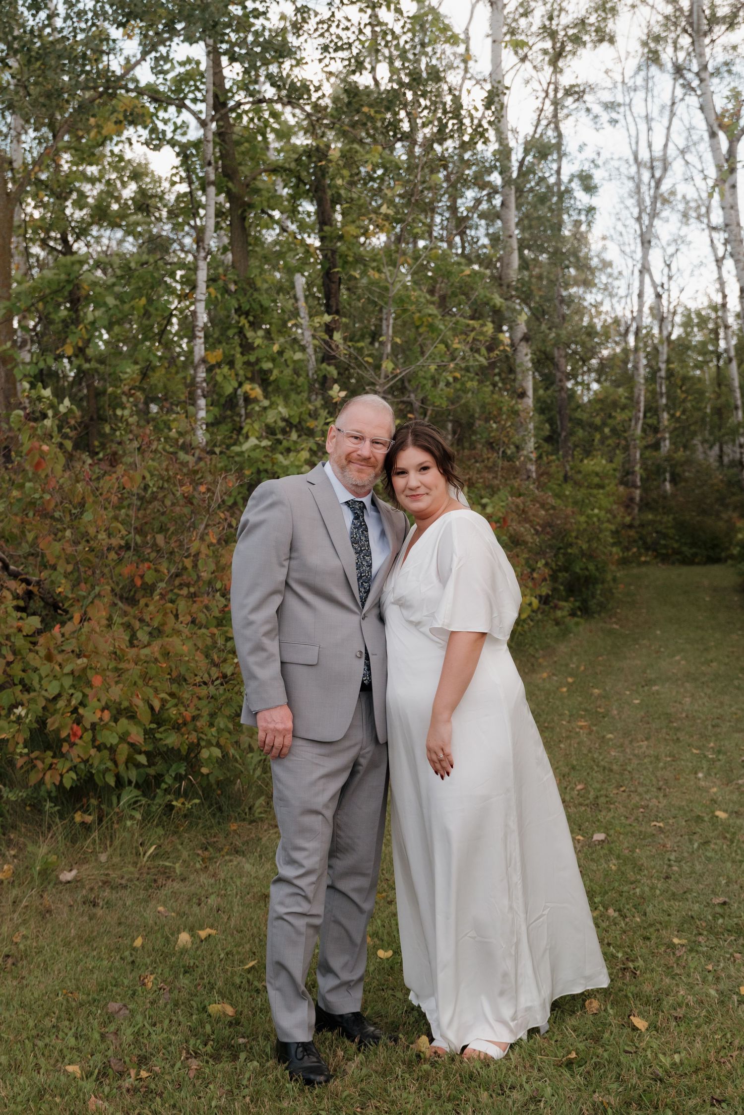 A couple in formal wedding attire poses outdoors in front of autumn trees and foliage.