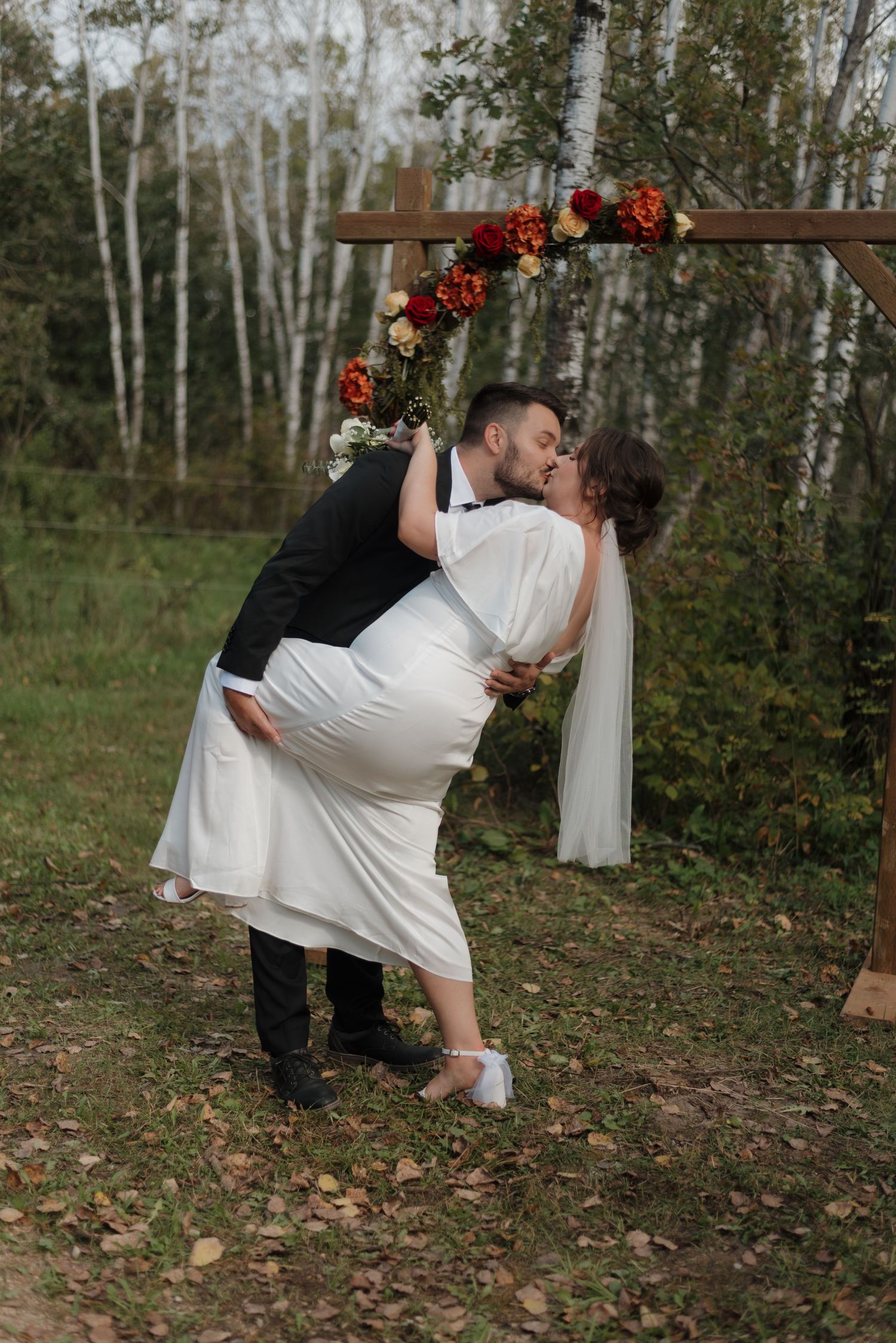 A romantic dip during an autumn woodland wedding ceremony under a wooden arch adorned with red flowers and white fabric.