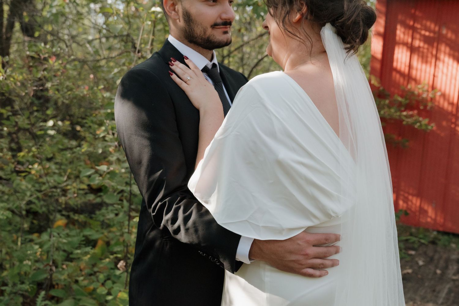 A romantic wedding moment between newlyweds in formal attire embracing outdoors near a rustic red wall and greenery.
