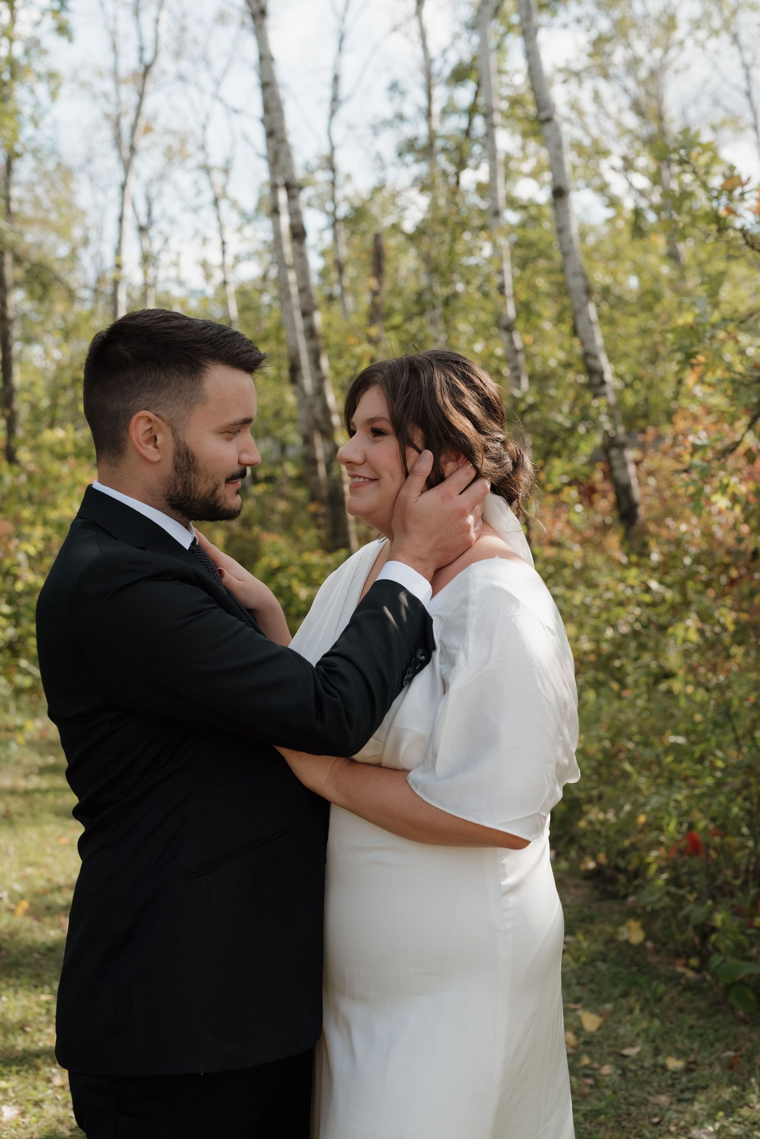 A tender wedding moment between newlyweds in white dress and black suit embracing in a sunlit forest setting.