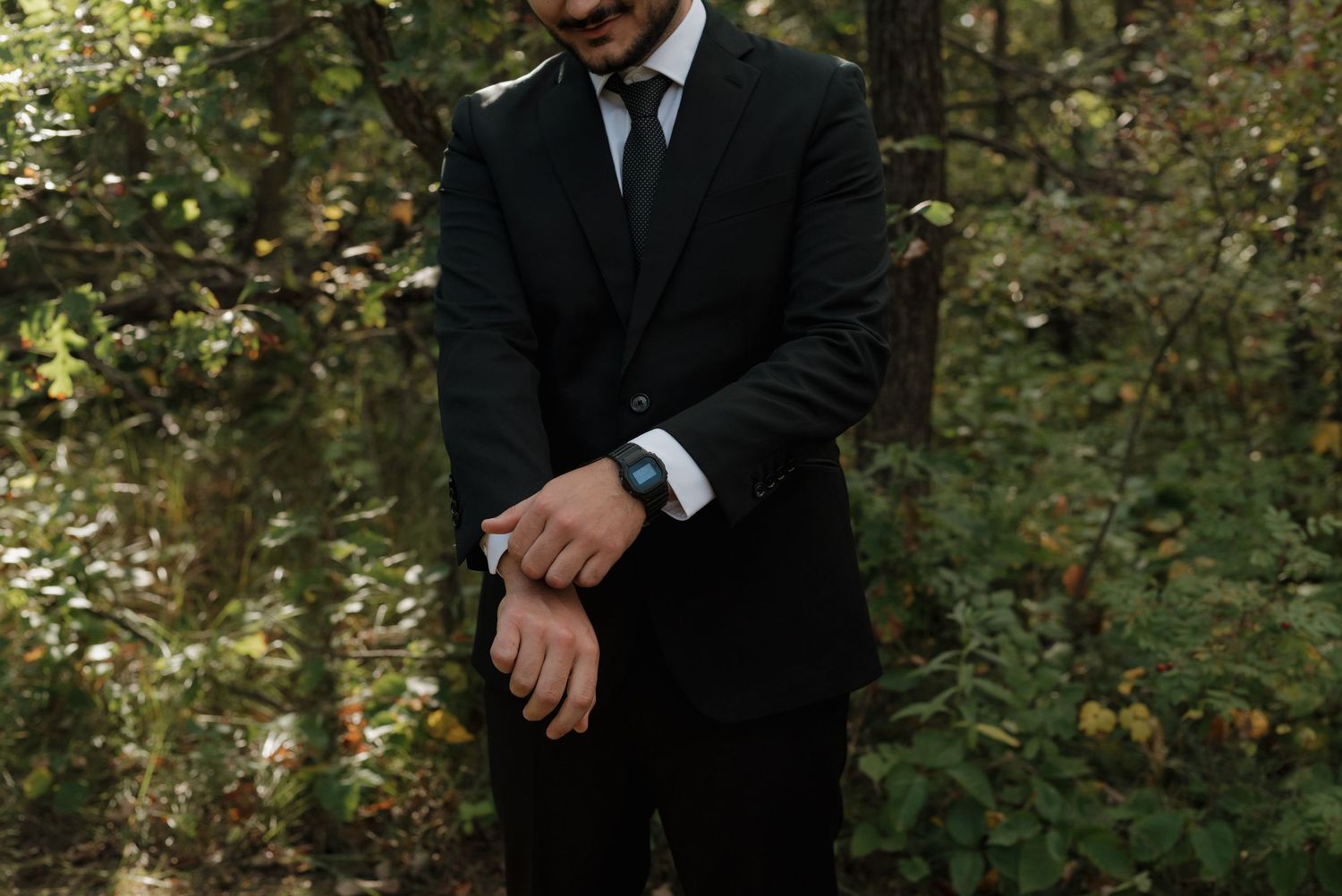 Person in black suit adjusts cufflinks while standing in a lush forest setting with dappled sunlight.