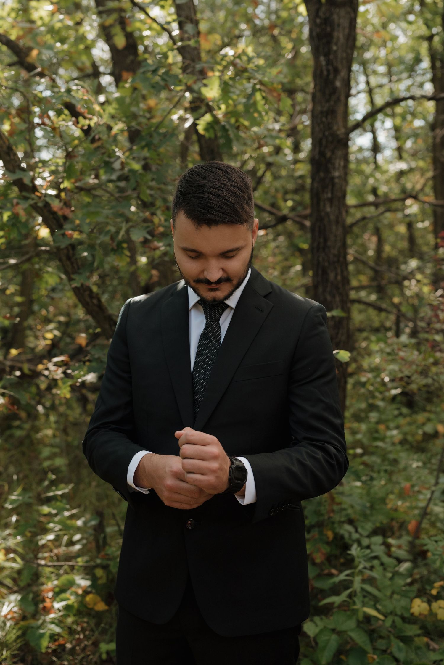 A groom in a black suit looks down thoughtfully during an outdoor wedding photo session.