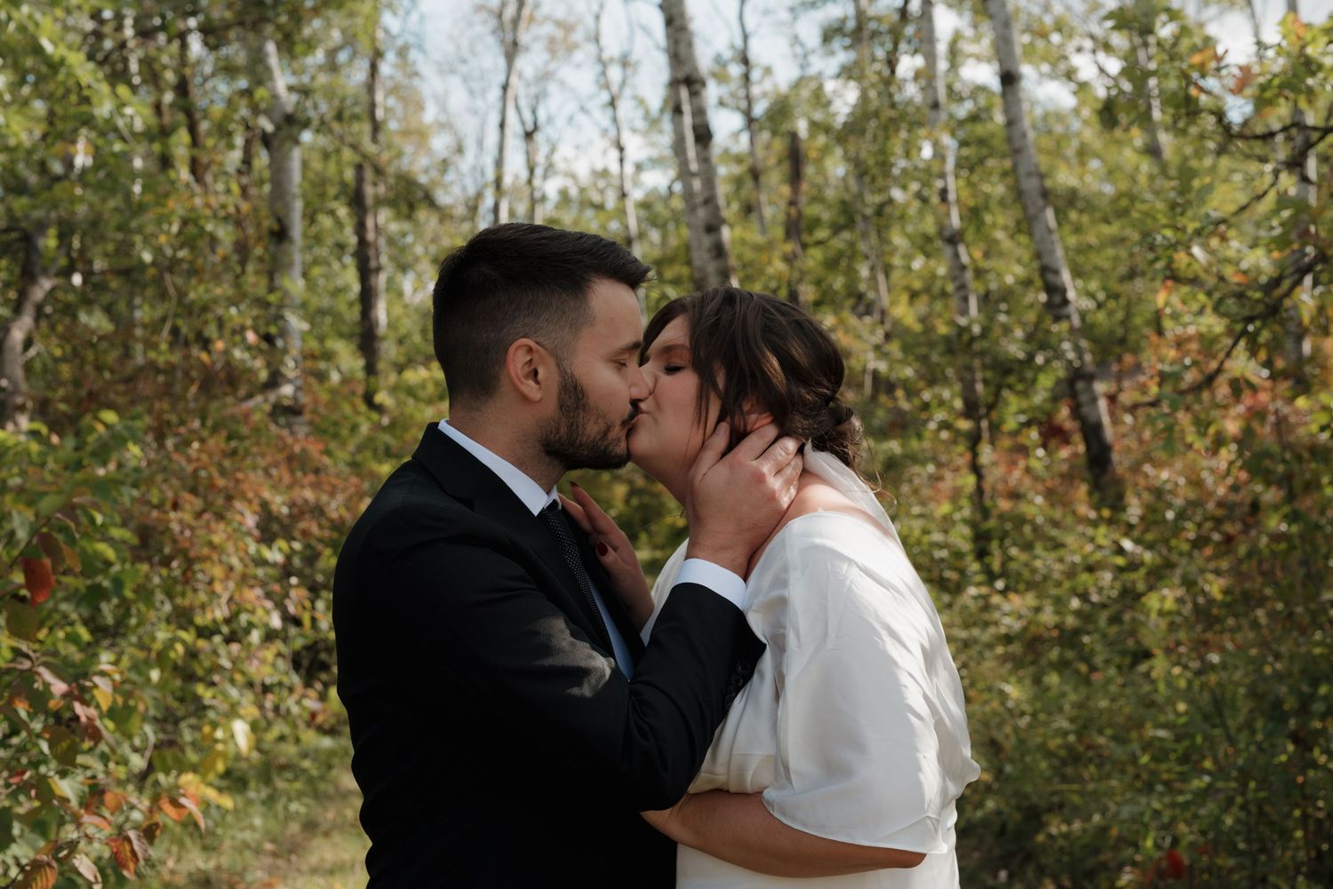 A romantic wedding couple shares a tender kiss in a beautiful autumn forest setting surrounded by fall foliage.