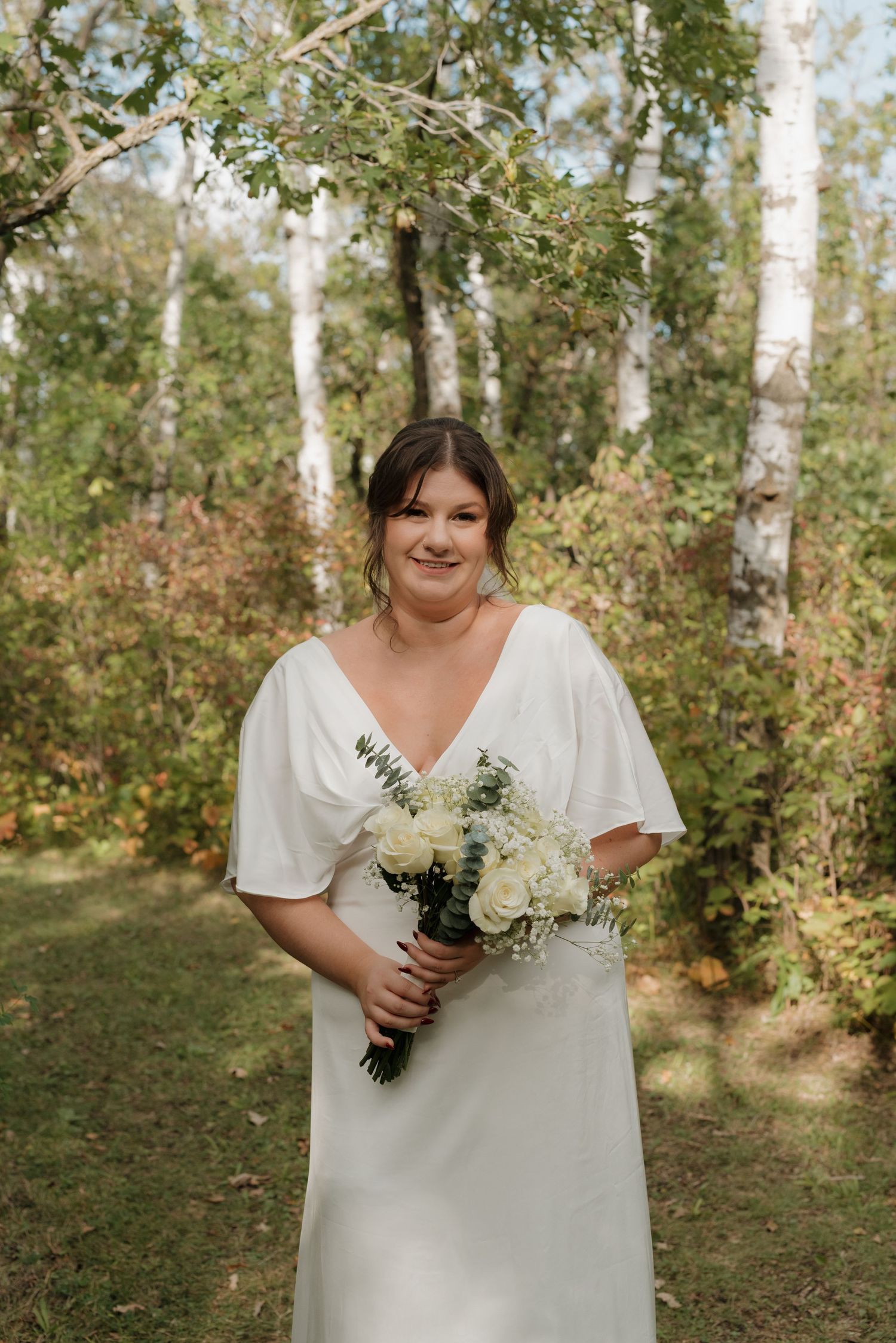 Someone in a white wedding dress holds a bouquet of white flowers while standing in a birch forest on a sunny day.