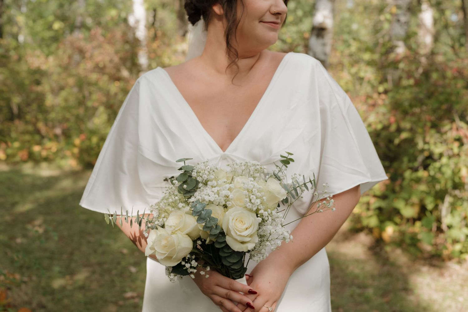 A bride in a flowing white v-neck dress holds an elegant bouquet of white roses and greenery in a garden setting.