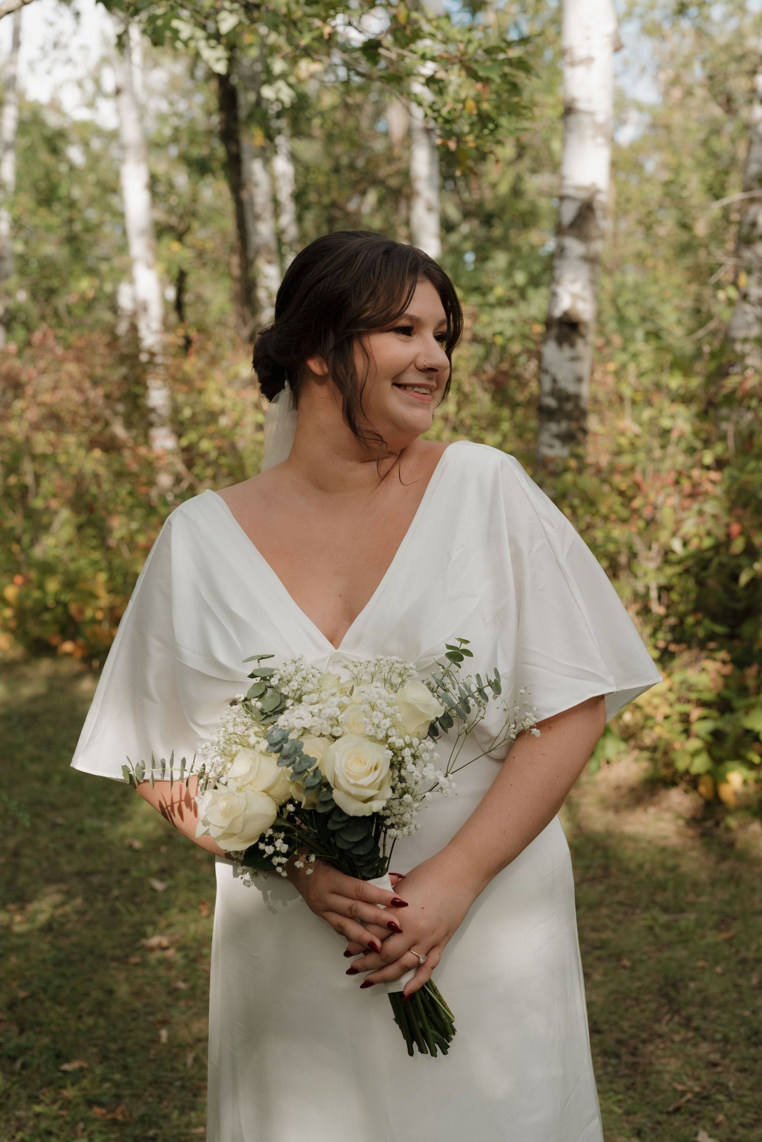 Bride in white dress holds a bouquet of white flowers while smiling in a wooded setting.