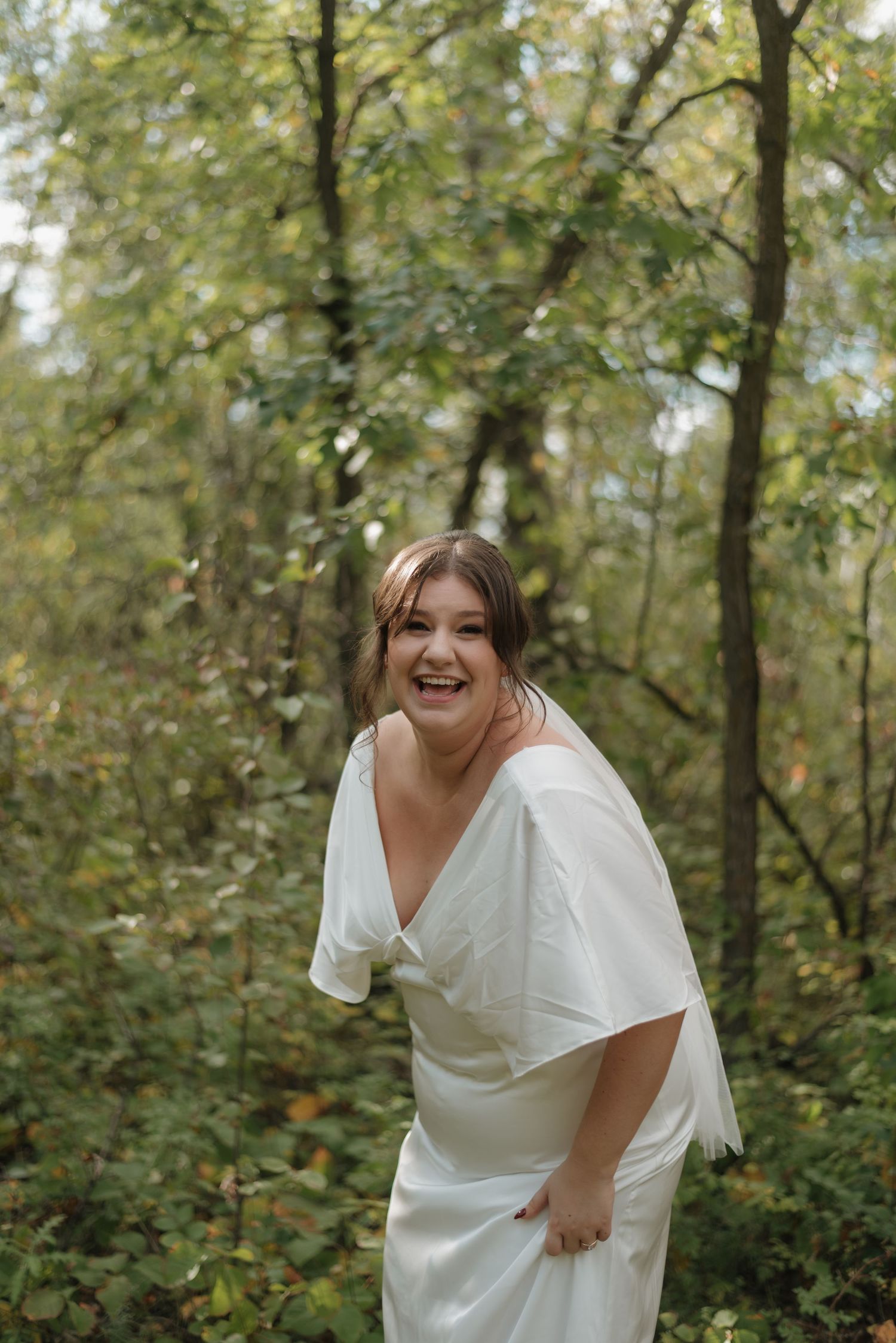 A joyful person laughs while wearing an elegant white dress during a forest photo shoot.