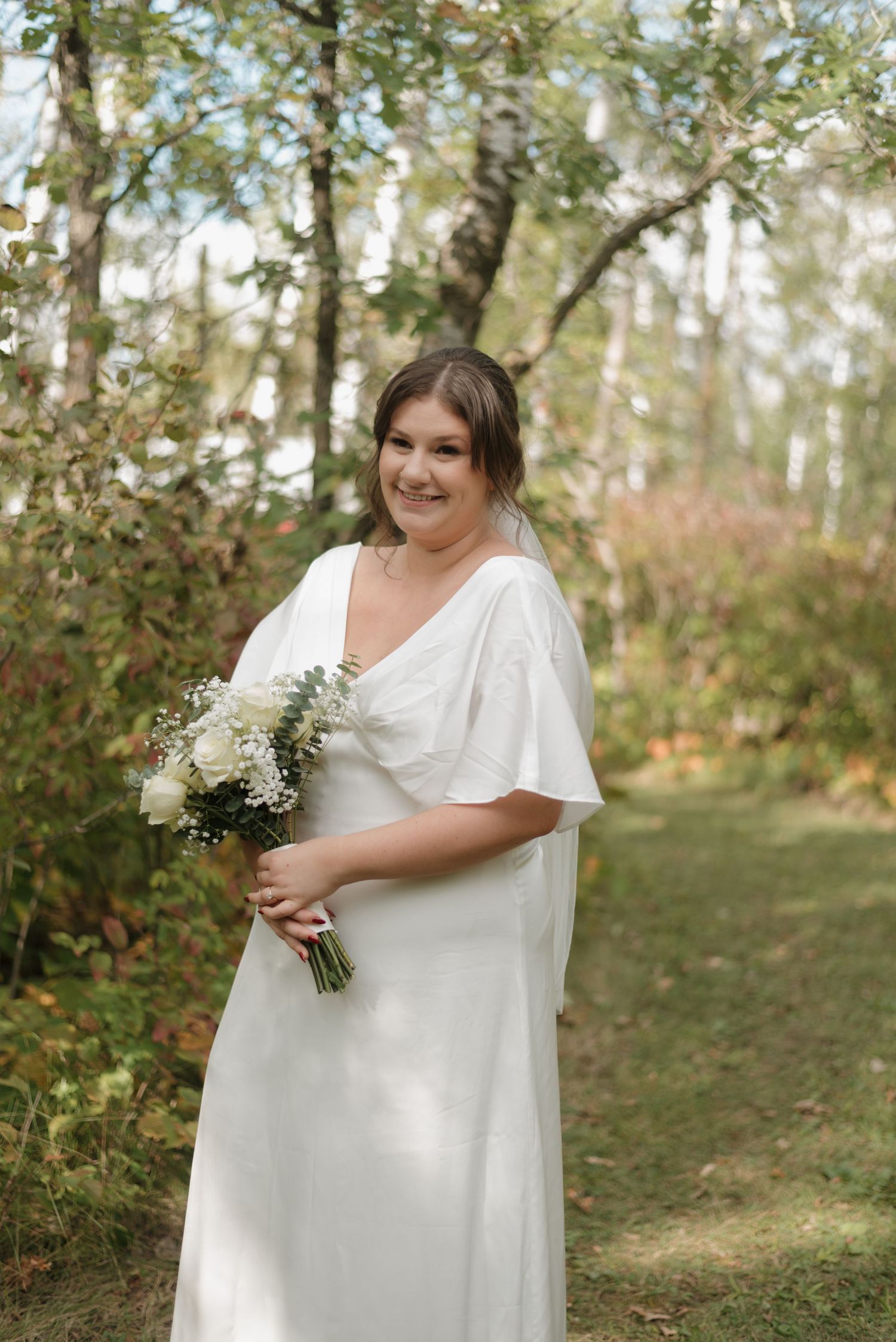 A bride in a flowing white dress with flutter sleeves holds a delicate white bouquet while standing in a wooded setting.