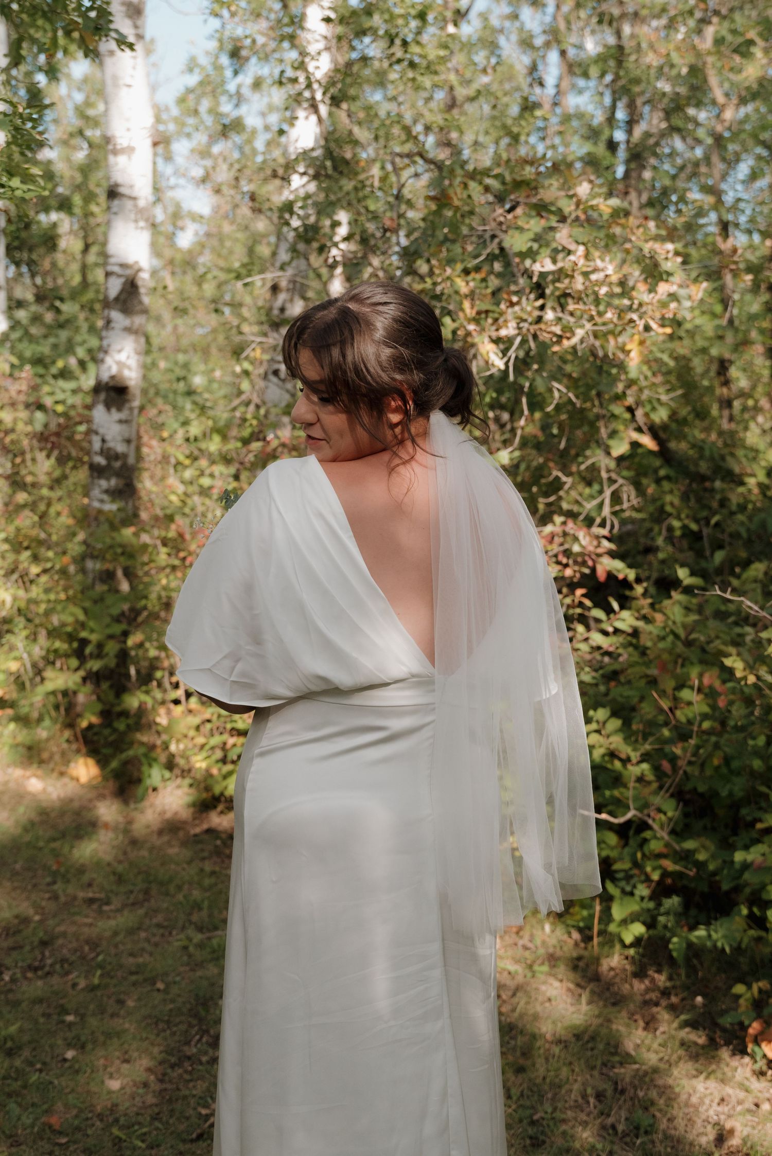 Bride in elegant white dress poses with her back turned in a serene forest setting.