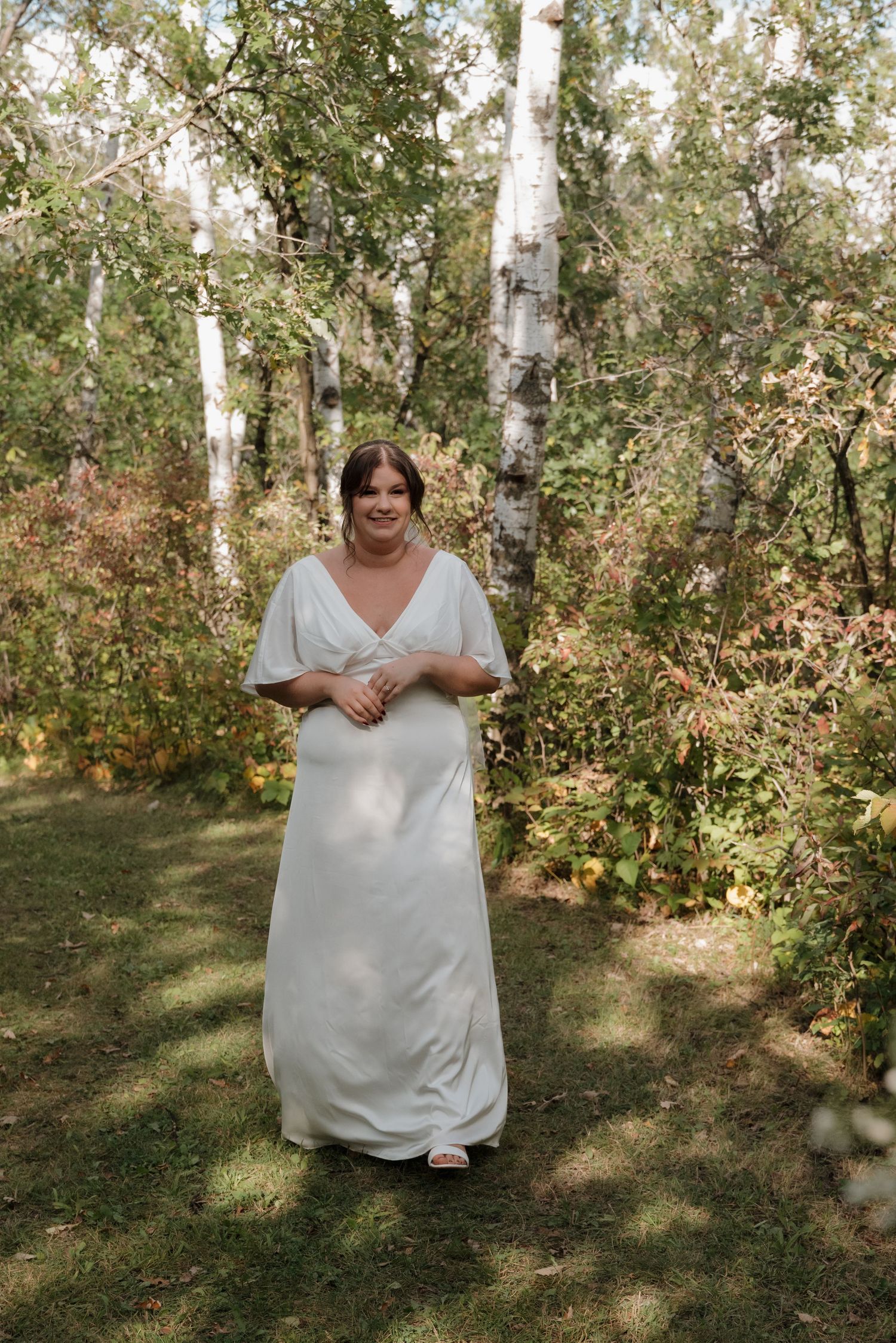 A radiant bride in a flowing white dress stands smiling in a serene birch forest setting.