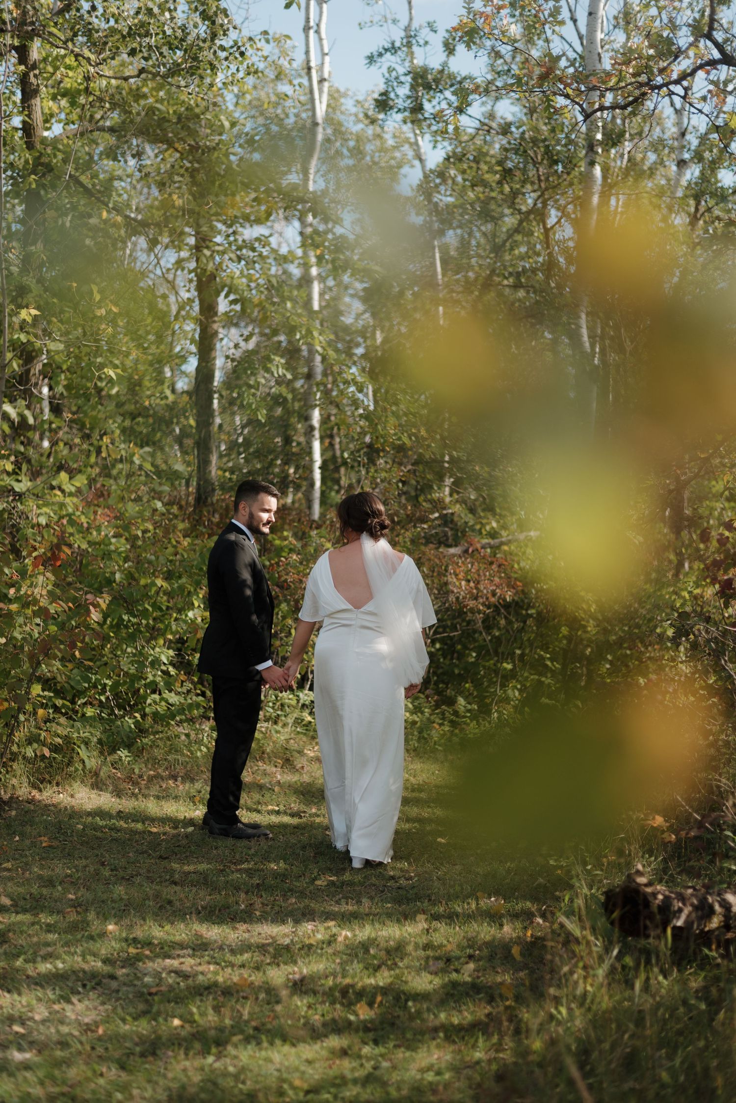 A couple in wedding attire stands together in a sun-dappled forest clearing on their special day.