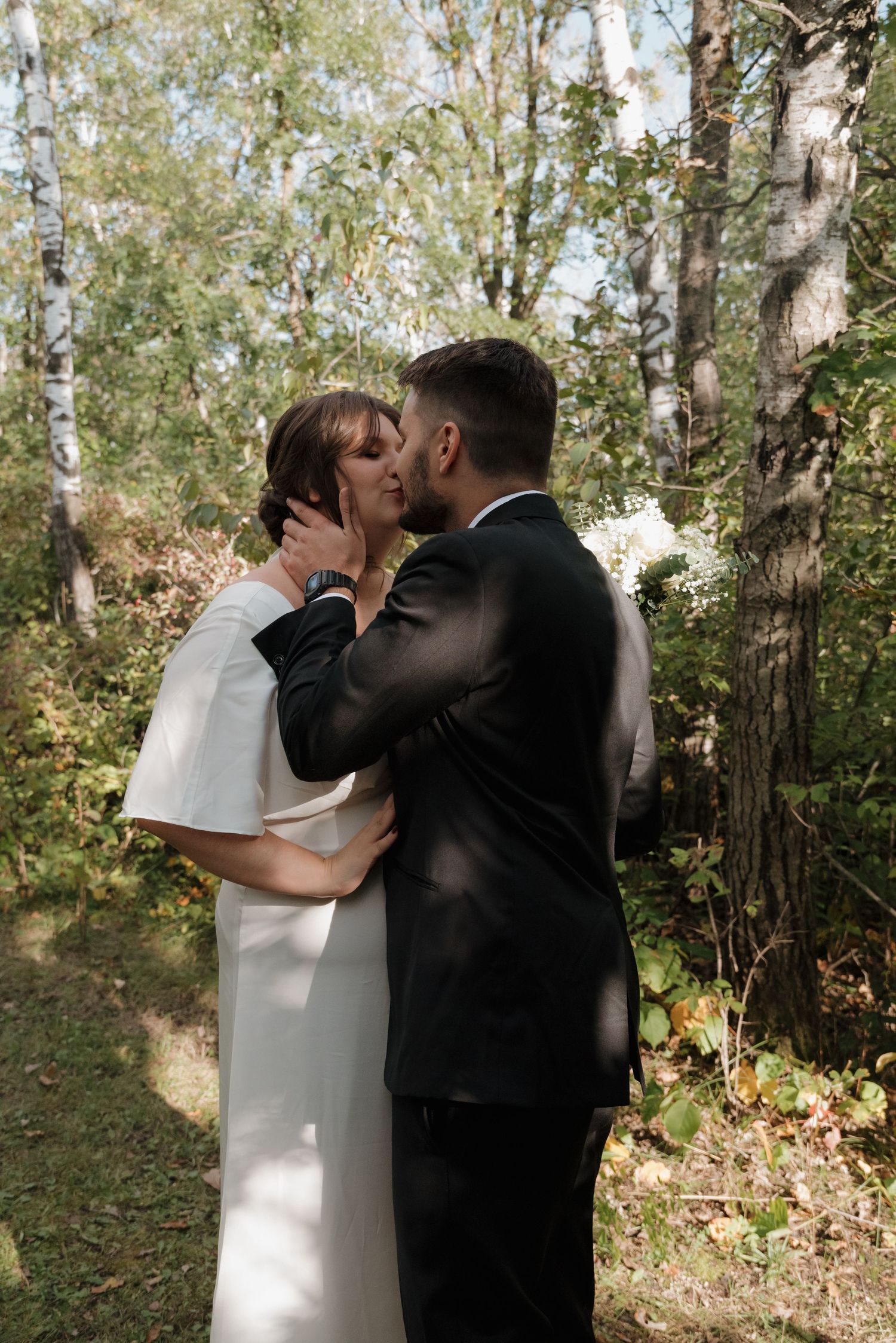 A romantic wedding moment captured between newlyweds sharing a tender kiss in a sunlit forest setting.