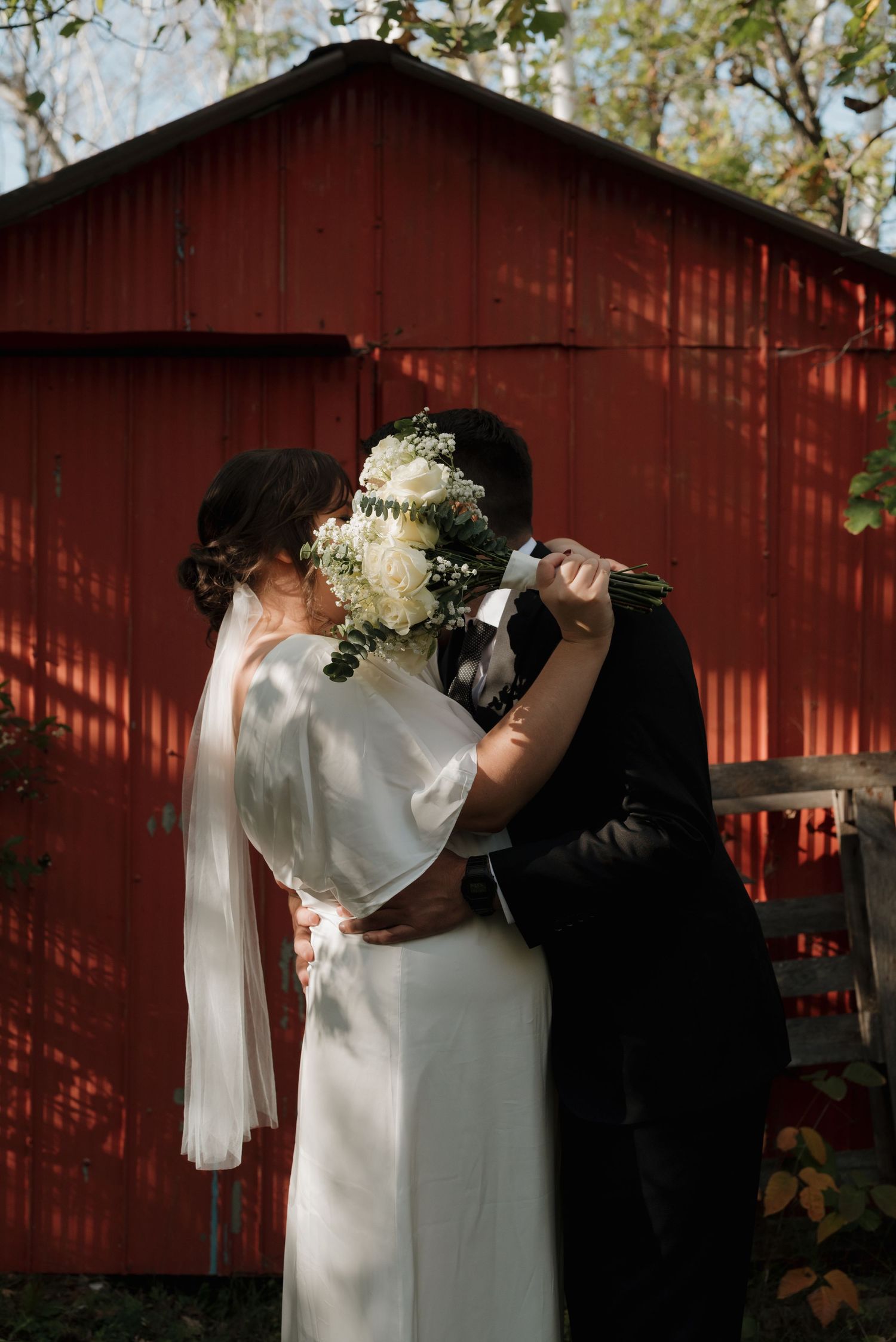 Couple sharing a romantic kiss behind a white bouquet in front of a rustic red barn on their wedding day.