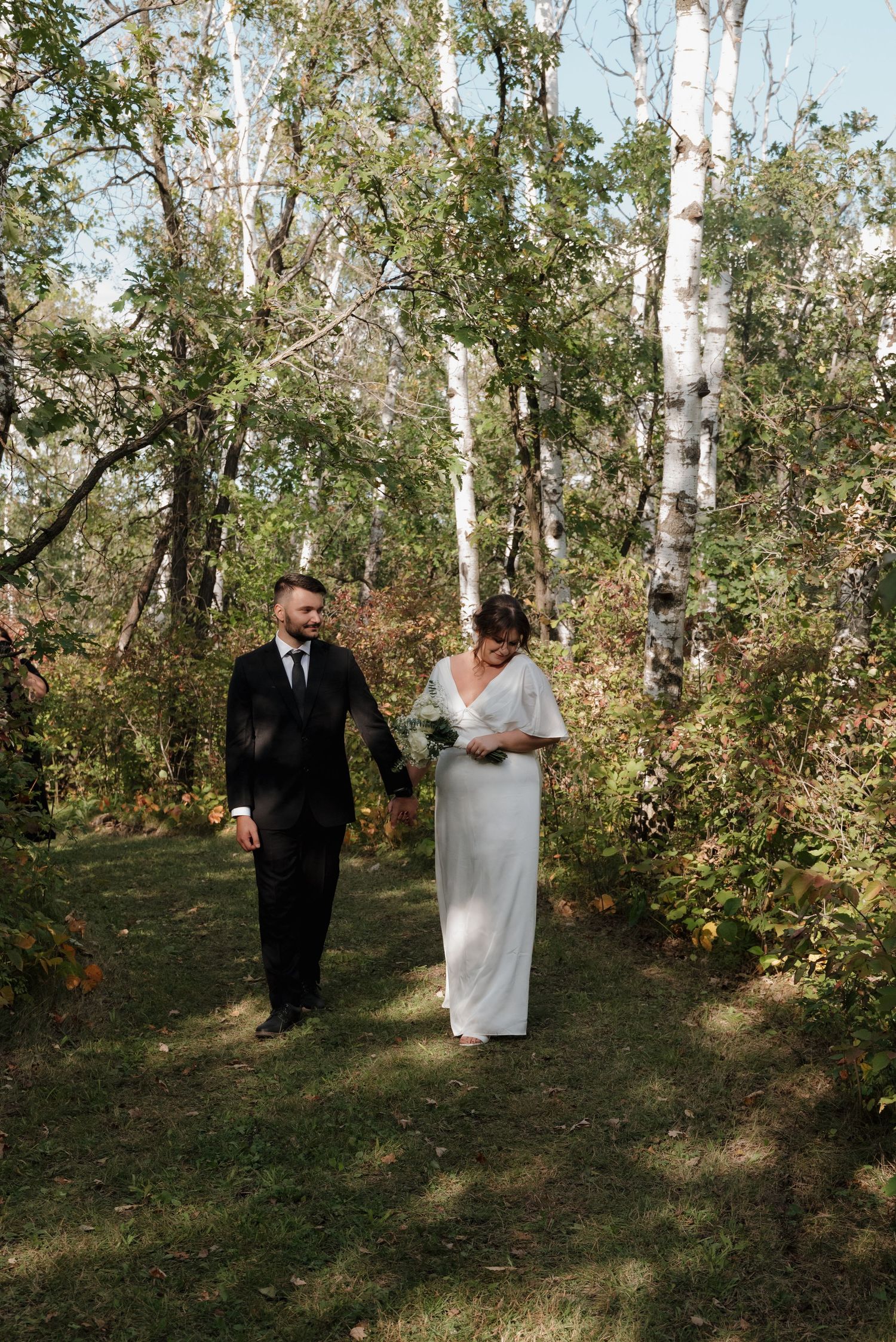 A couple walks together along a wooded path with birch trees and greenery on a sunny autumn day.