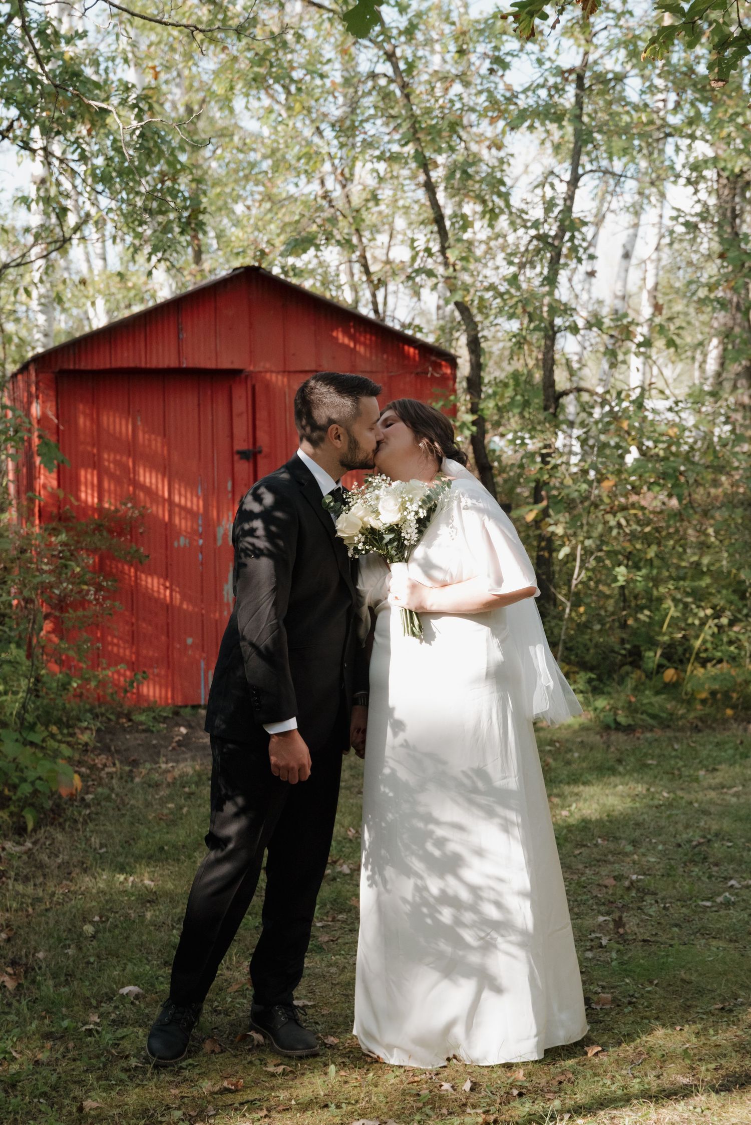 A rustic wedding couple shares a romantic moment in front of a red barn surrounded by birch trees and fall foliage.
