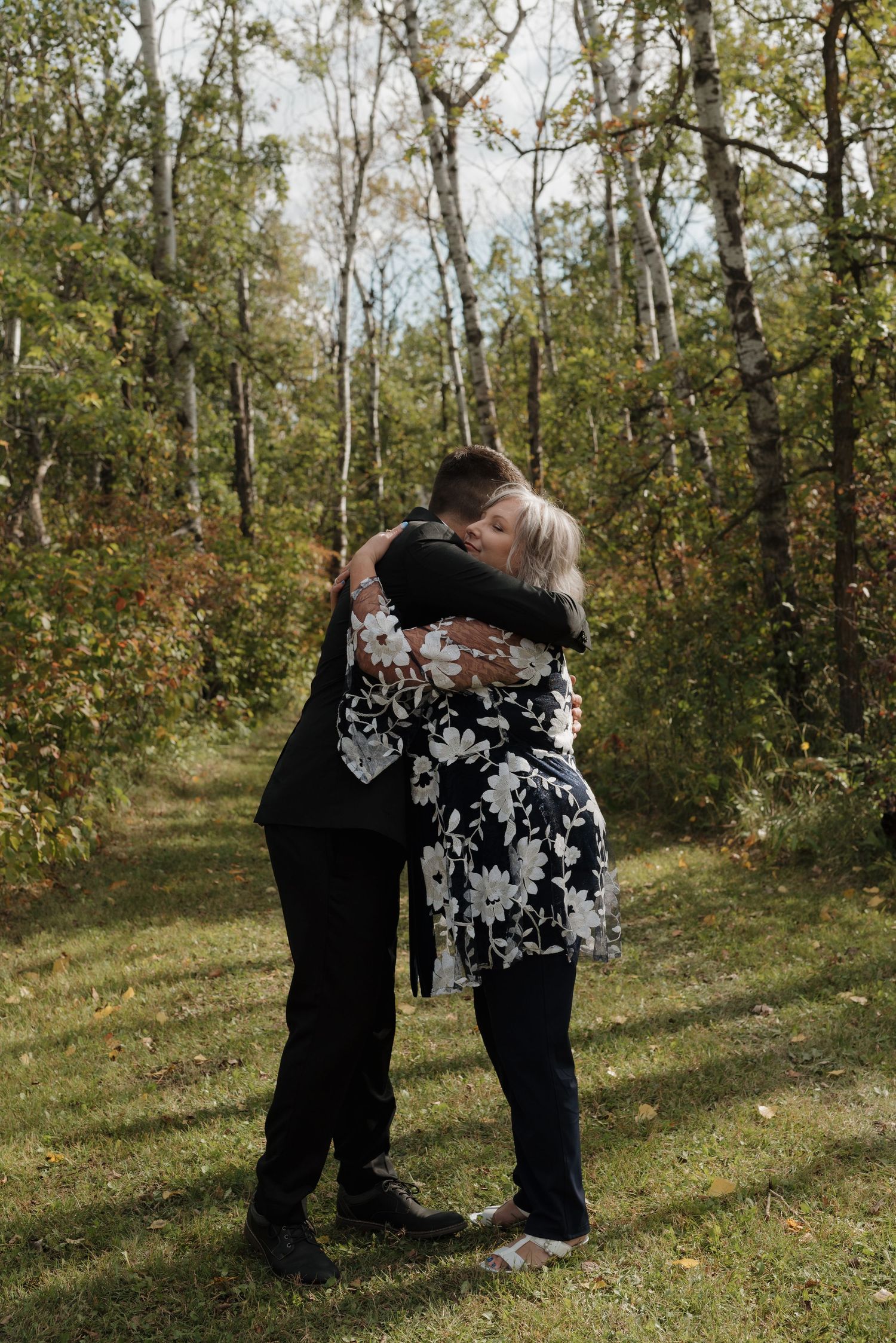 Two people share a tender embrace while standing in a sunlit forest clearing surrounded by autumn trees.
