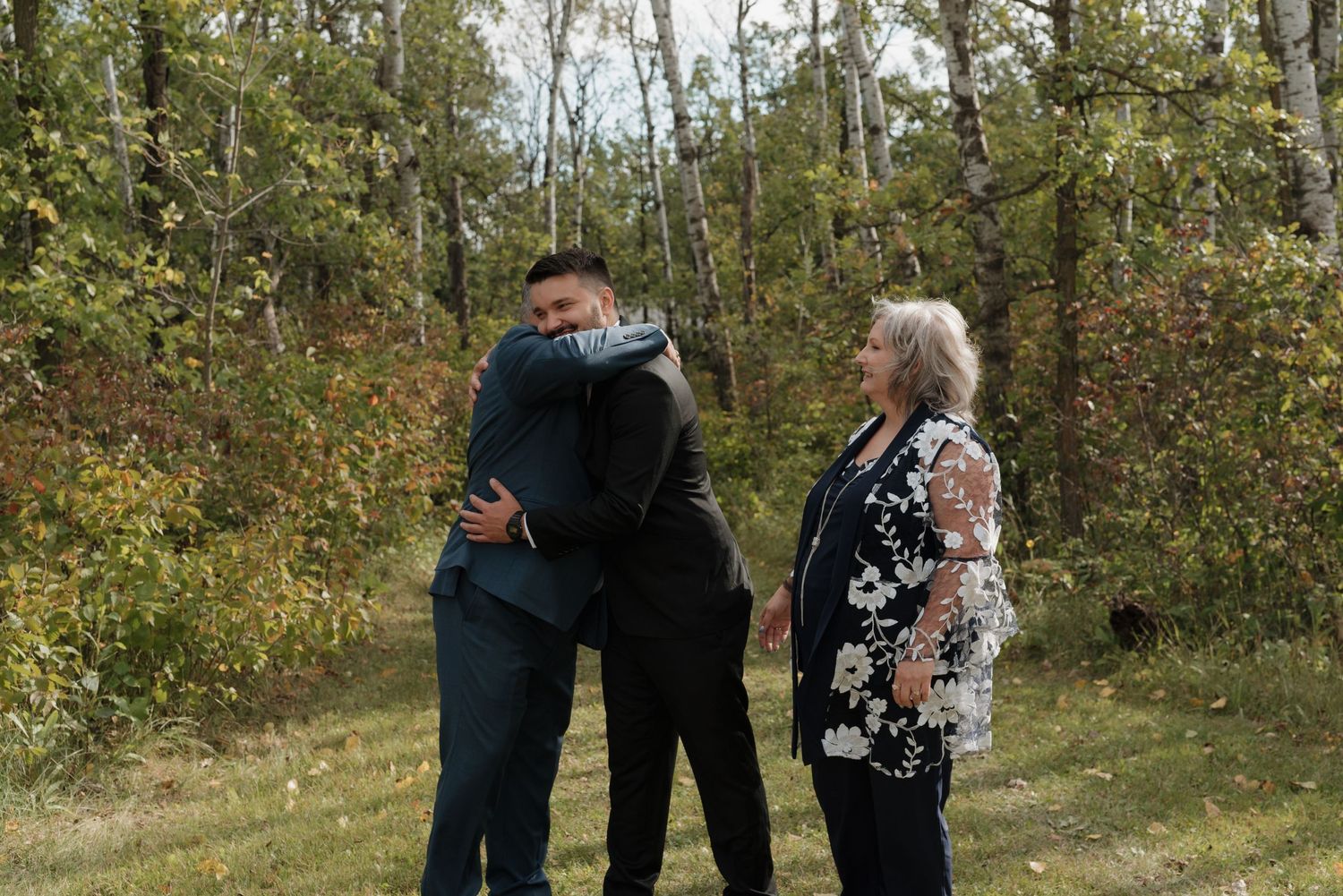 A small group embraces during an emotional outdoor gathering surrounded by autumn trees and foliage.