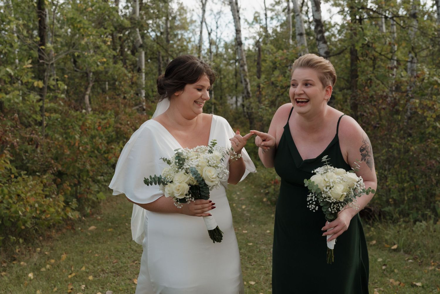 Two people laugh together while holding white floral bouquets outdoors in a wooded setting.