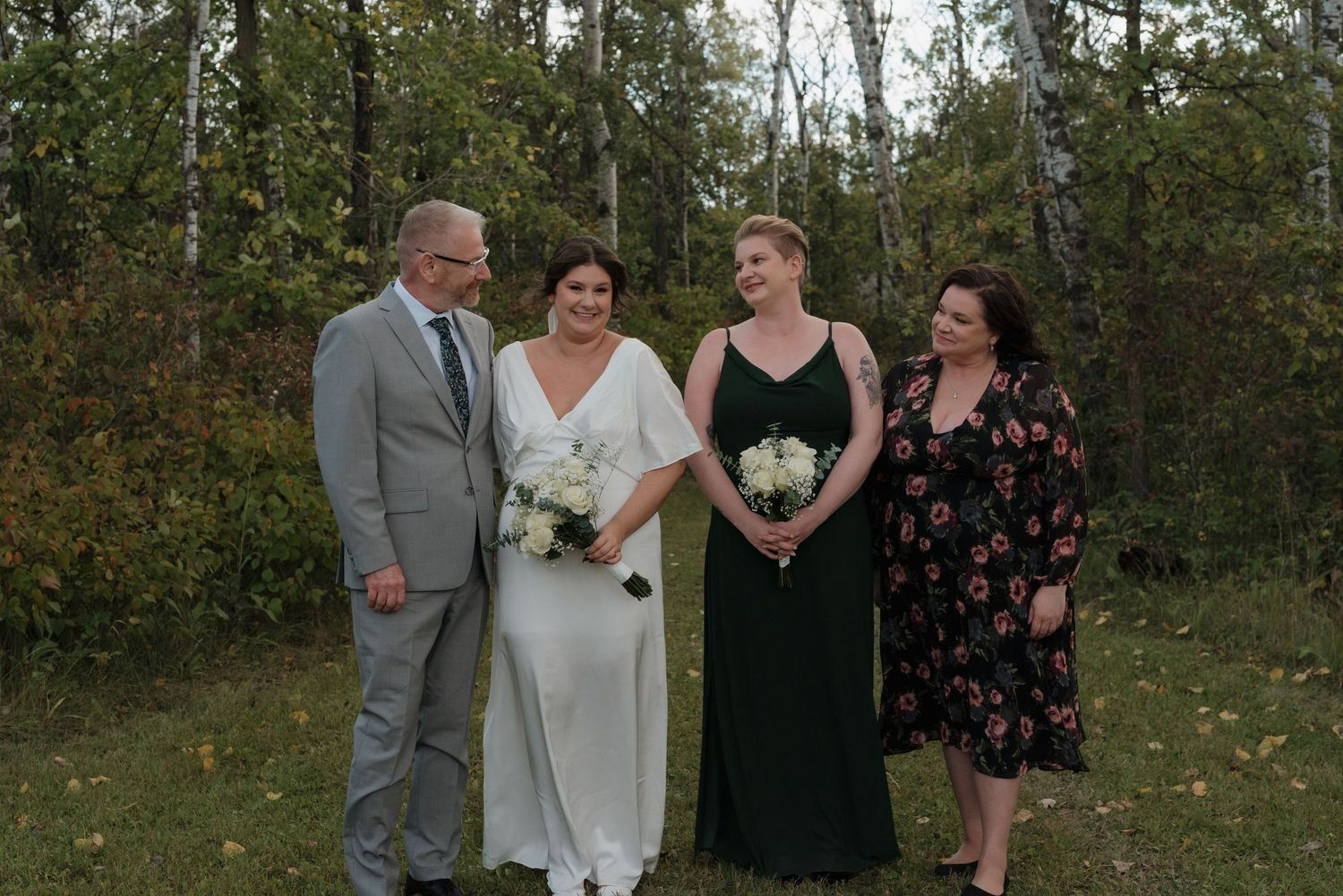 Wedding party standing together outdoors in formal attire against green foliage backdrop.