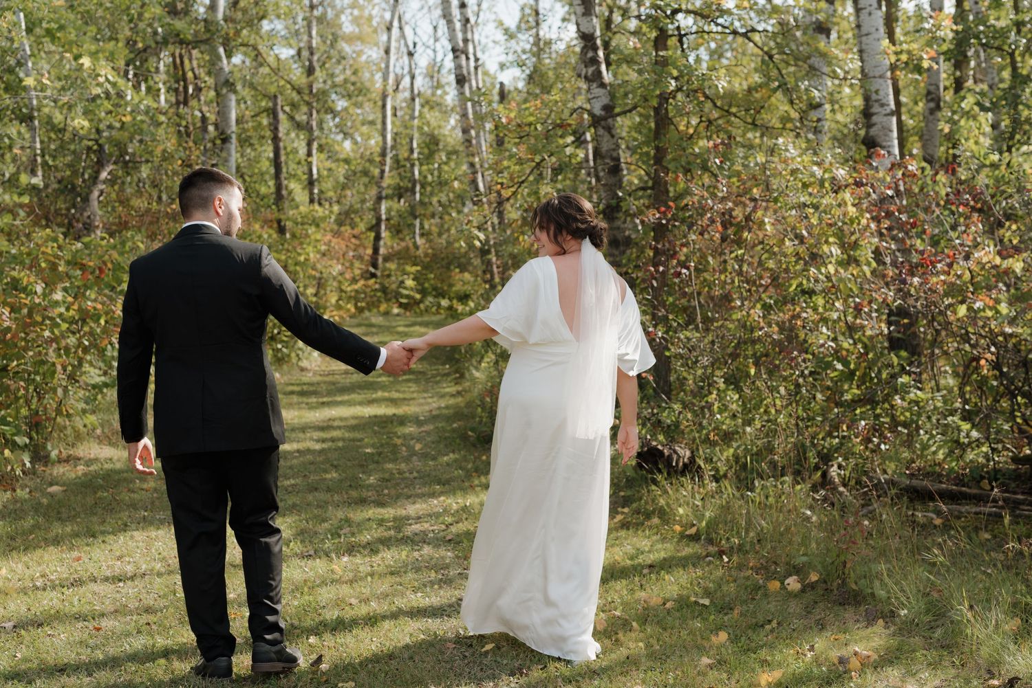 A bride and groom hold hands while walking on a wooded path during their outdoor wedding photoshoot.