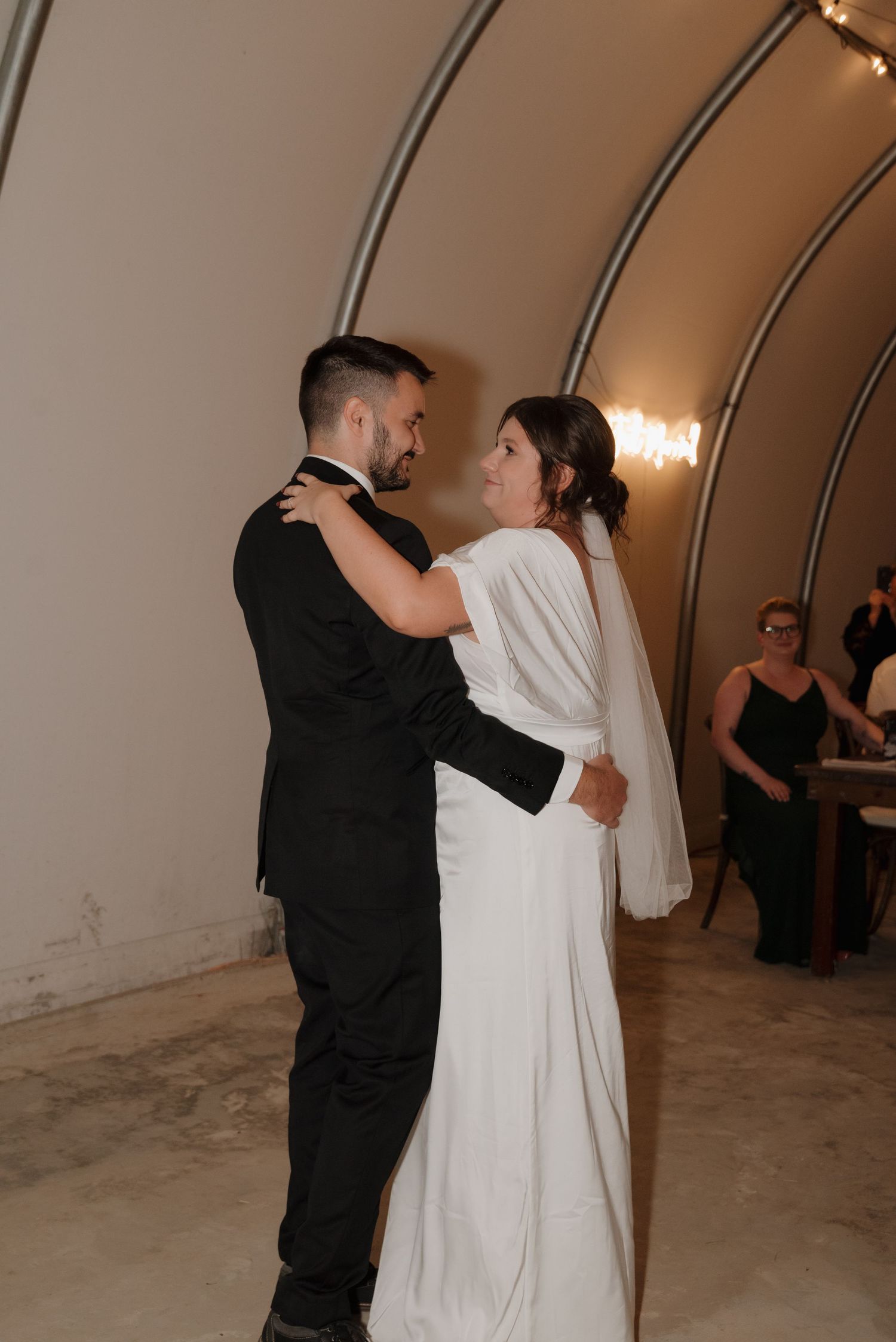 A couple shares their first dance at their wedding reception in an elegant venue with curved walls and soft lighting.