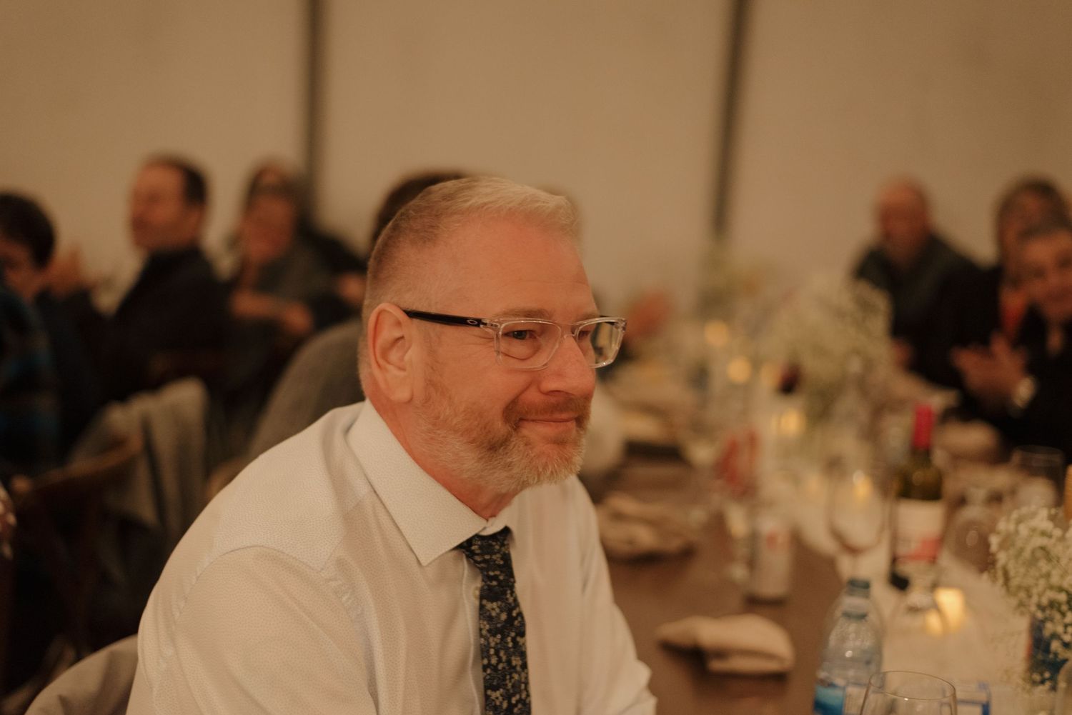 A person in glasses and white dress shirt with tie smiles warmly while seated at a formal dining event.