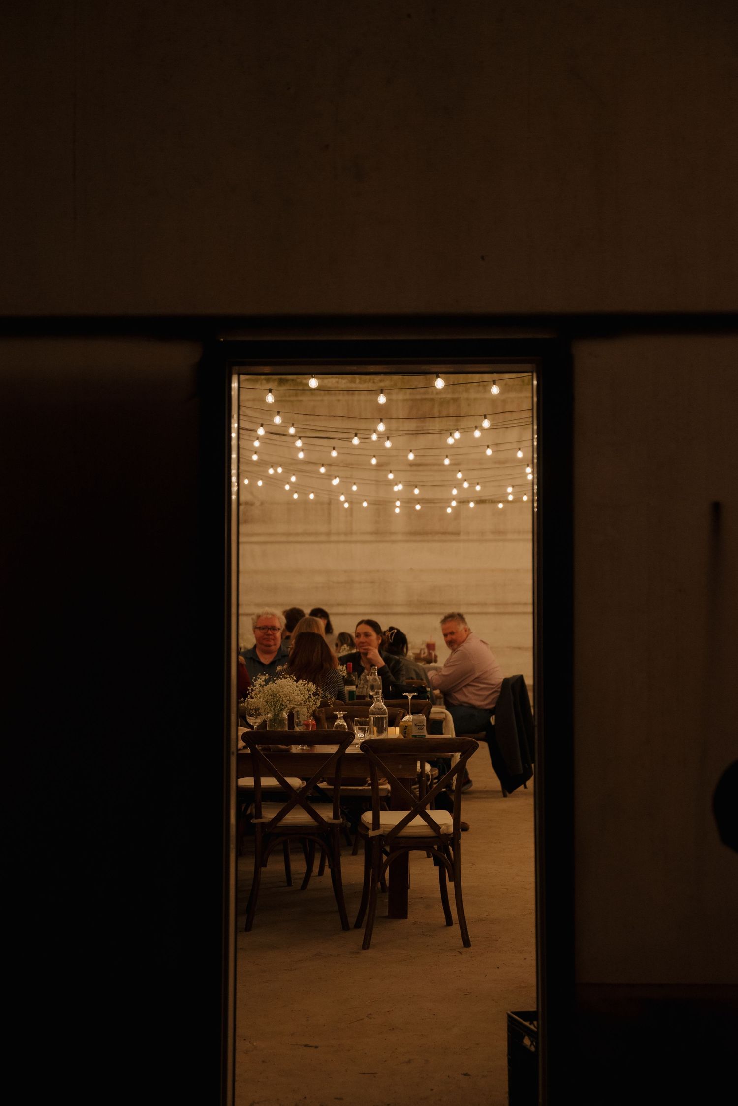 A warm glow illuminates a dining room through a doorway, with string lights hung above and diners gathered at wooden tables.