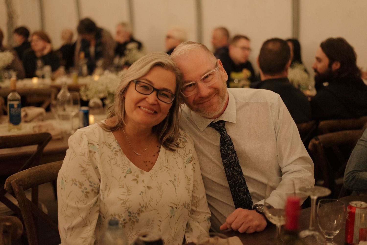 Wedding guests smile warmly while seated at reception tables with cozy ambient lighting.