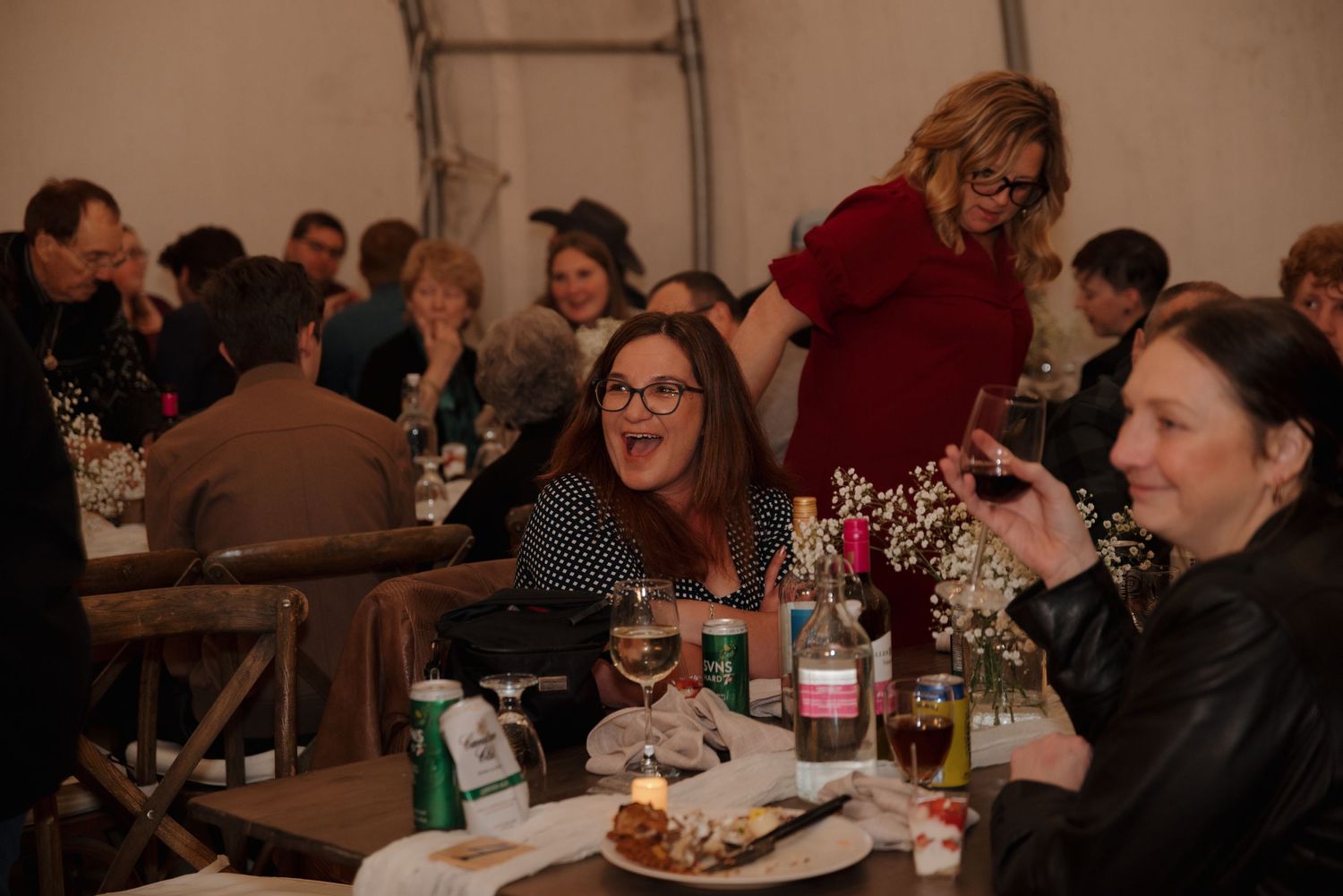 Wedding guests enjoy a festive reception dinner with wine and table decorations in a tented venue.