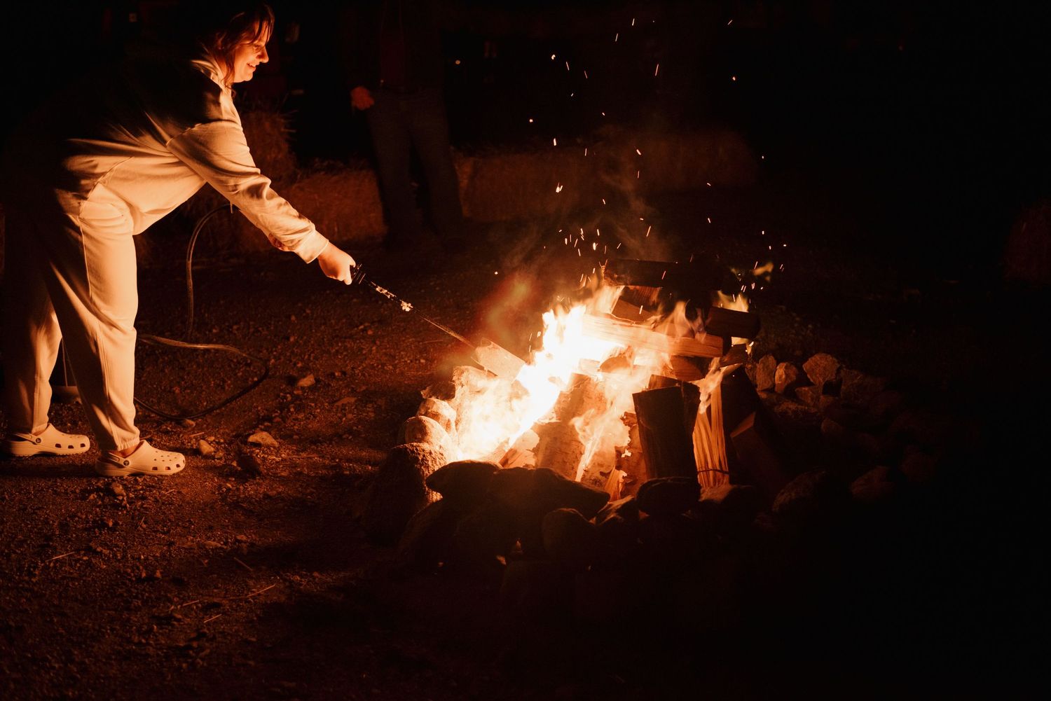 Person stoking a nighttime bonfire with sparks flying into the dark sky.