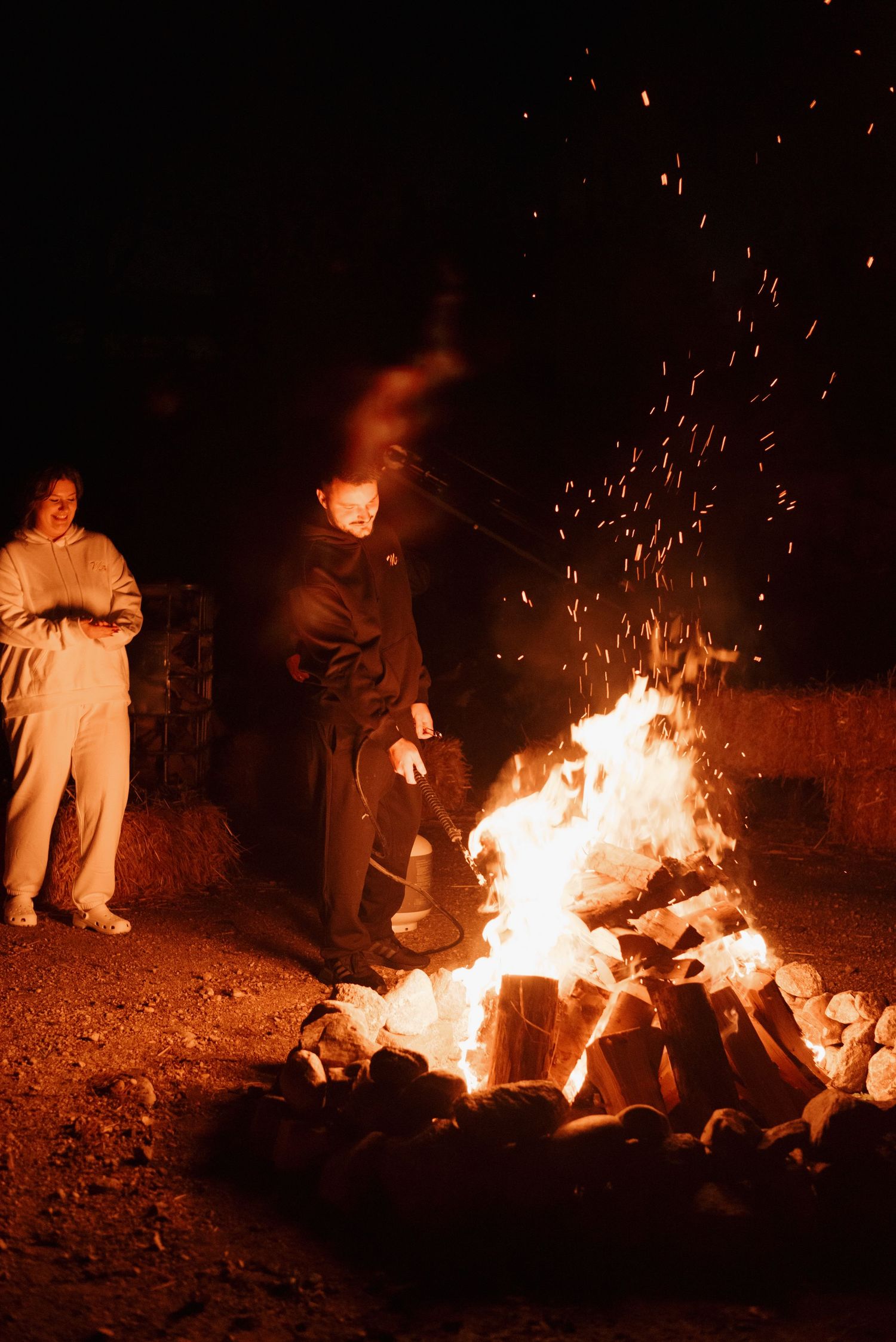 People stand next to a large bonfire with sparks flying into the dark night sky as flames illuminate the scene.