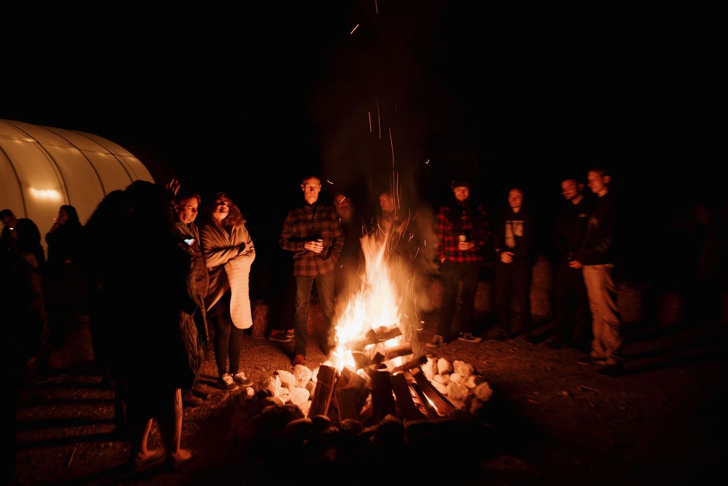 Wedding guests gather around a large bonfire at night creating a cozy outdoor atmosphere.