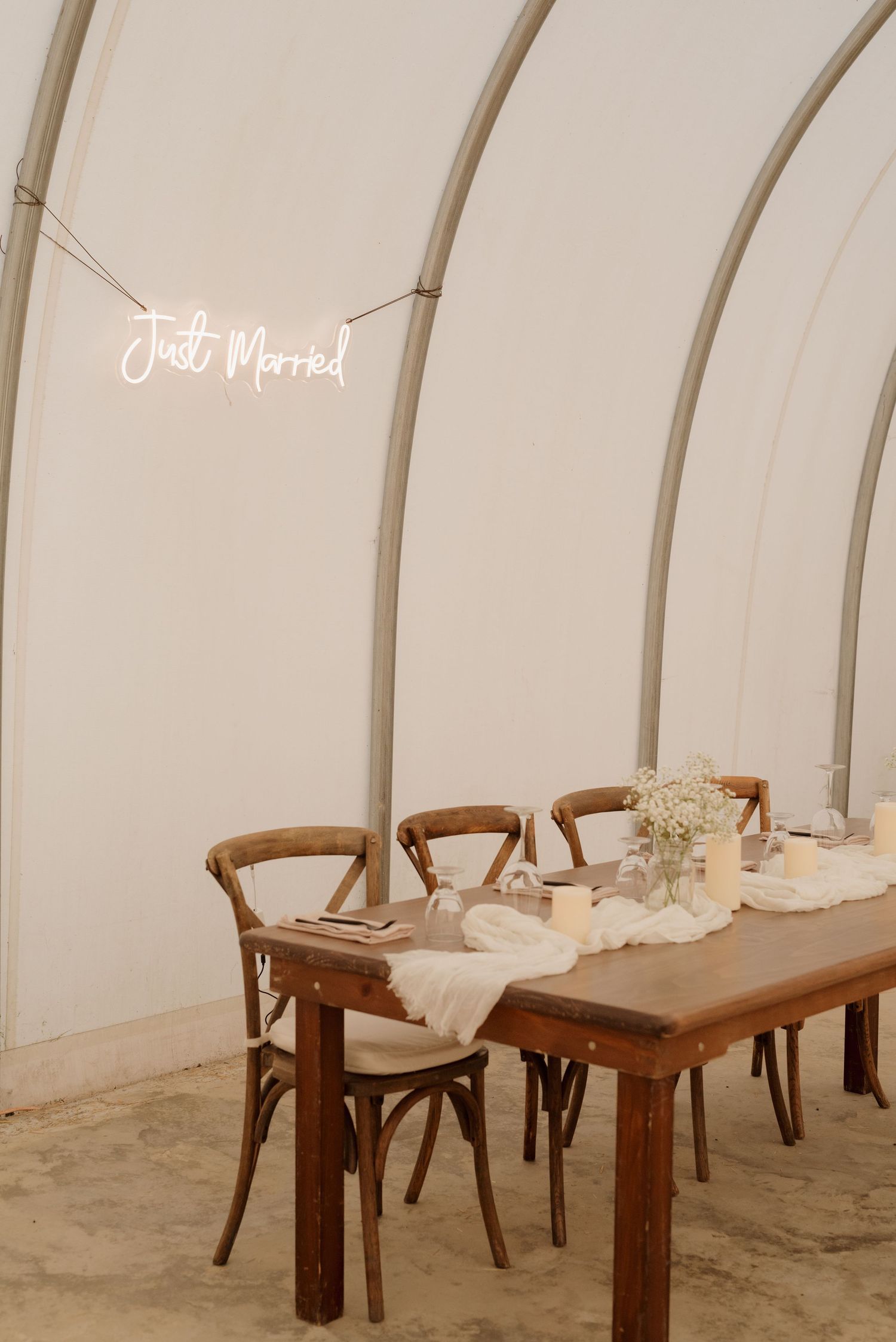 Rustic wedding table setup with wooden chairs and neon 'Just Married' sign in a white tent structure.