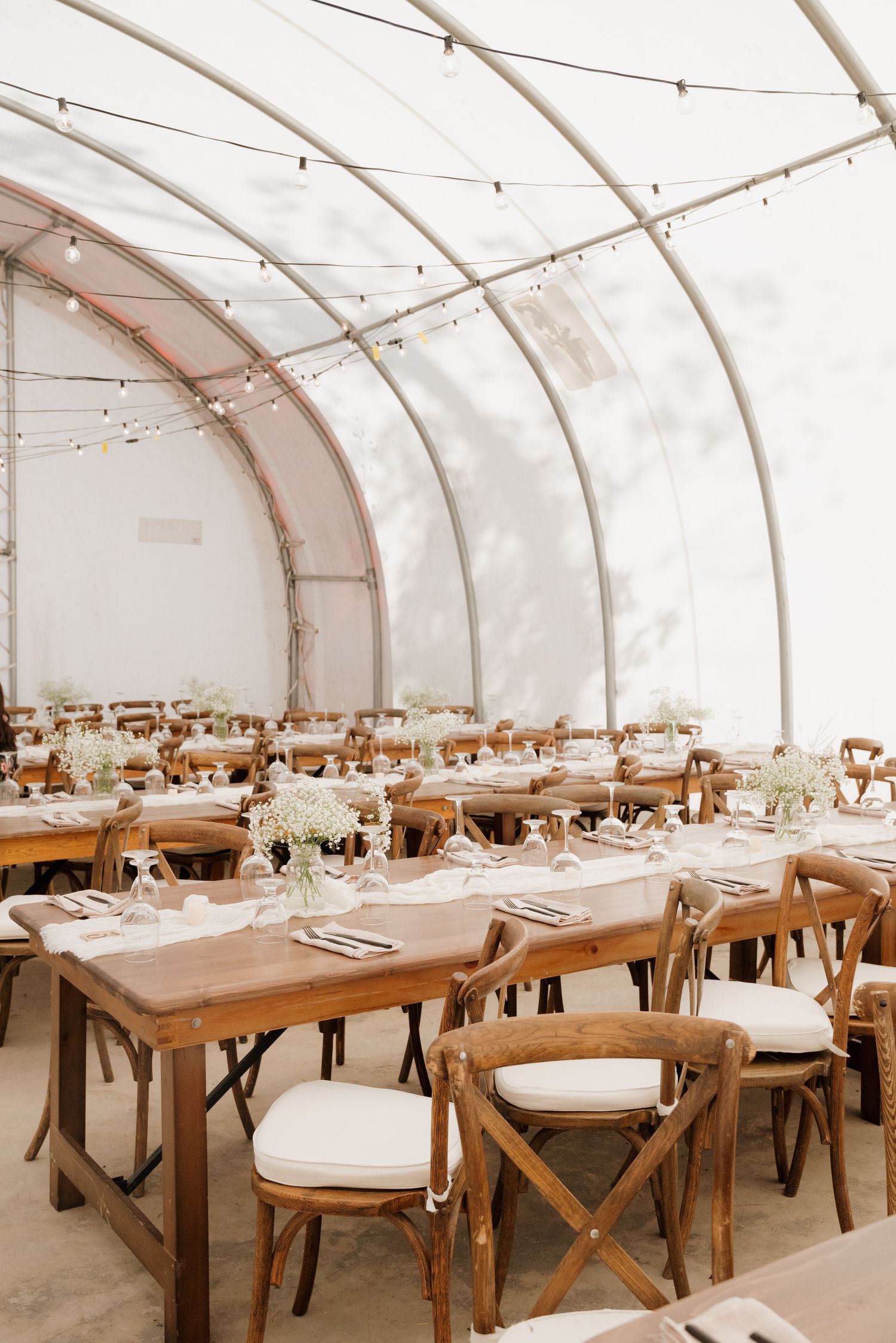 Elegant wedding reception setup in a white tent with rustic wooden farm tables and crossback chairs.