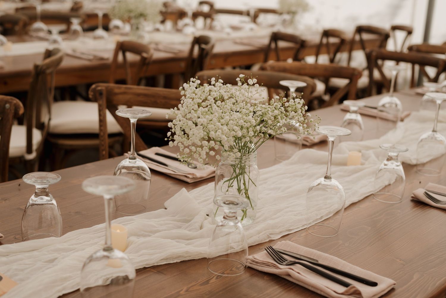 Elegant rustic wedding table setting with baby's breath centerpieces on white table runners and wooden chairs.