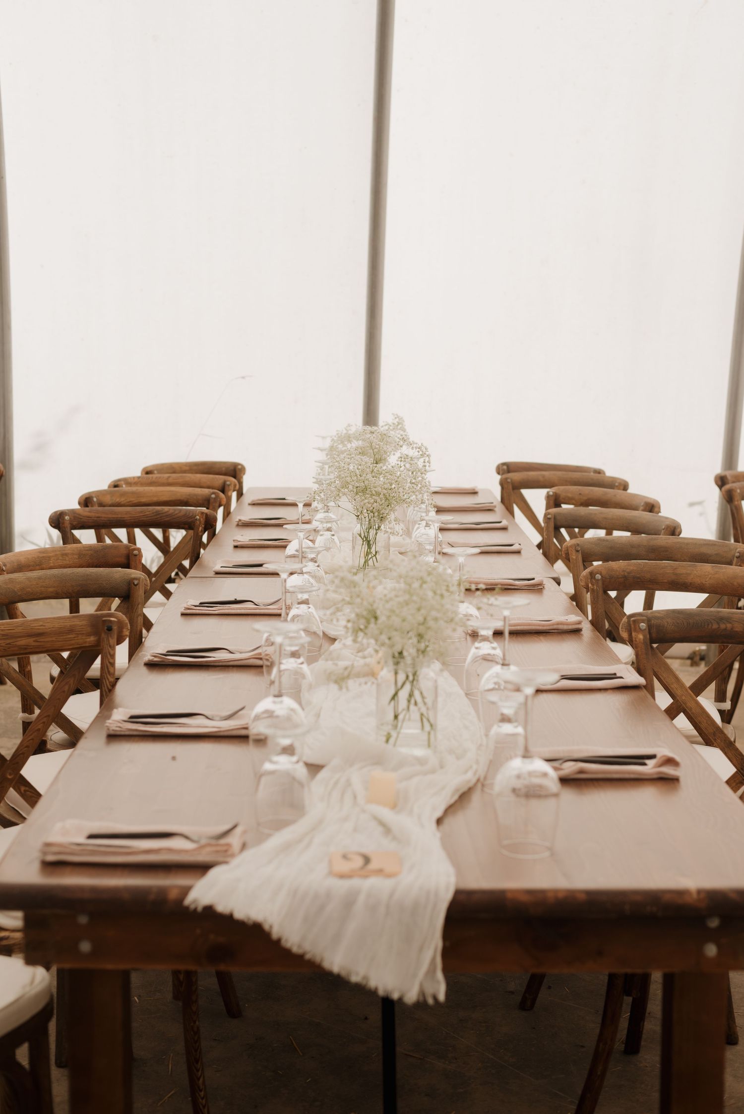 Elegant table setting with white fabric runner, baby's breath centerpieces and cross-back chairs in white tent venue.