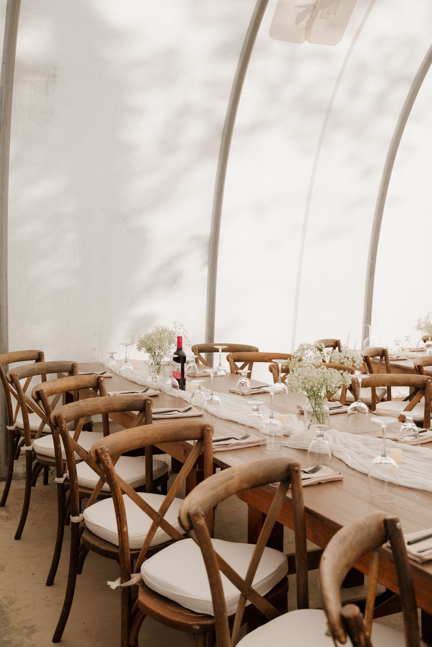 Long wooden dining tables with white cushioned crossback chairs set under a white tent canopy.