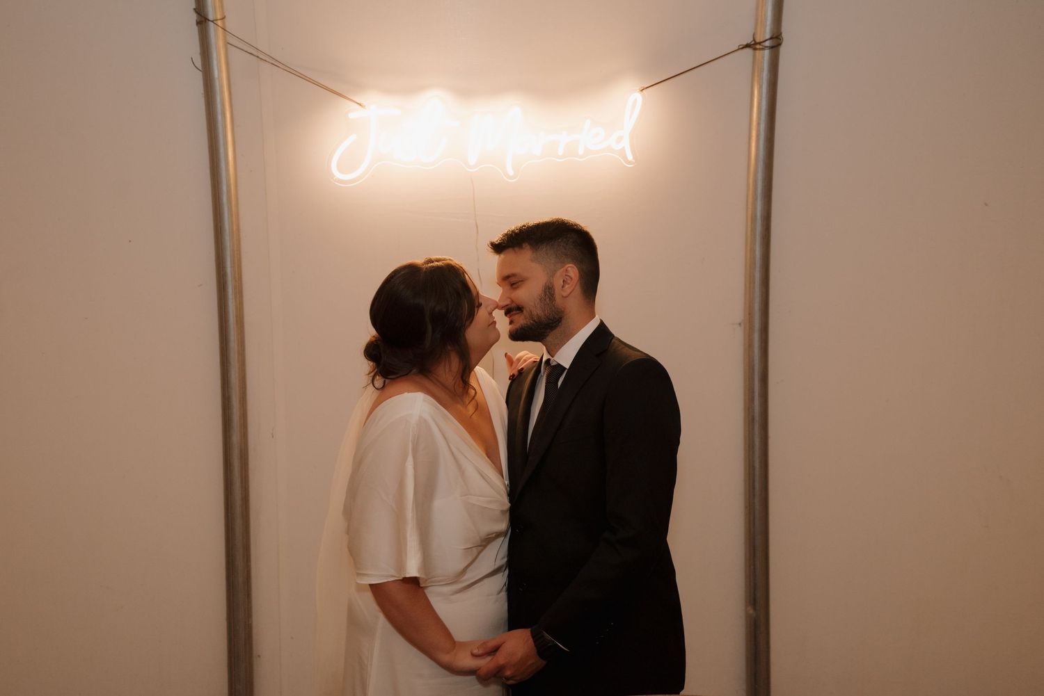 Couple shares intimate moment under glowing 'til death' neon sign at wedding venue.