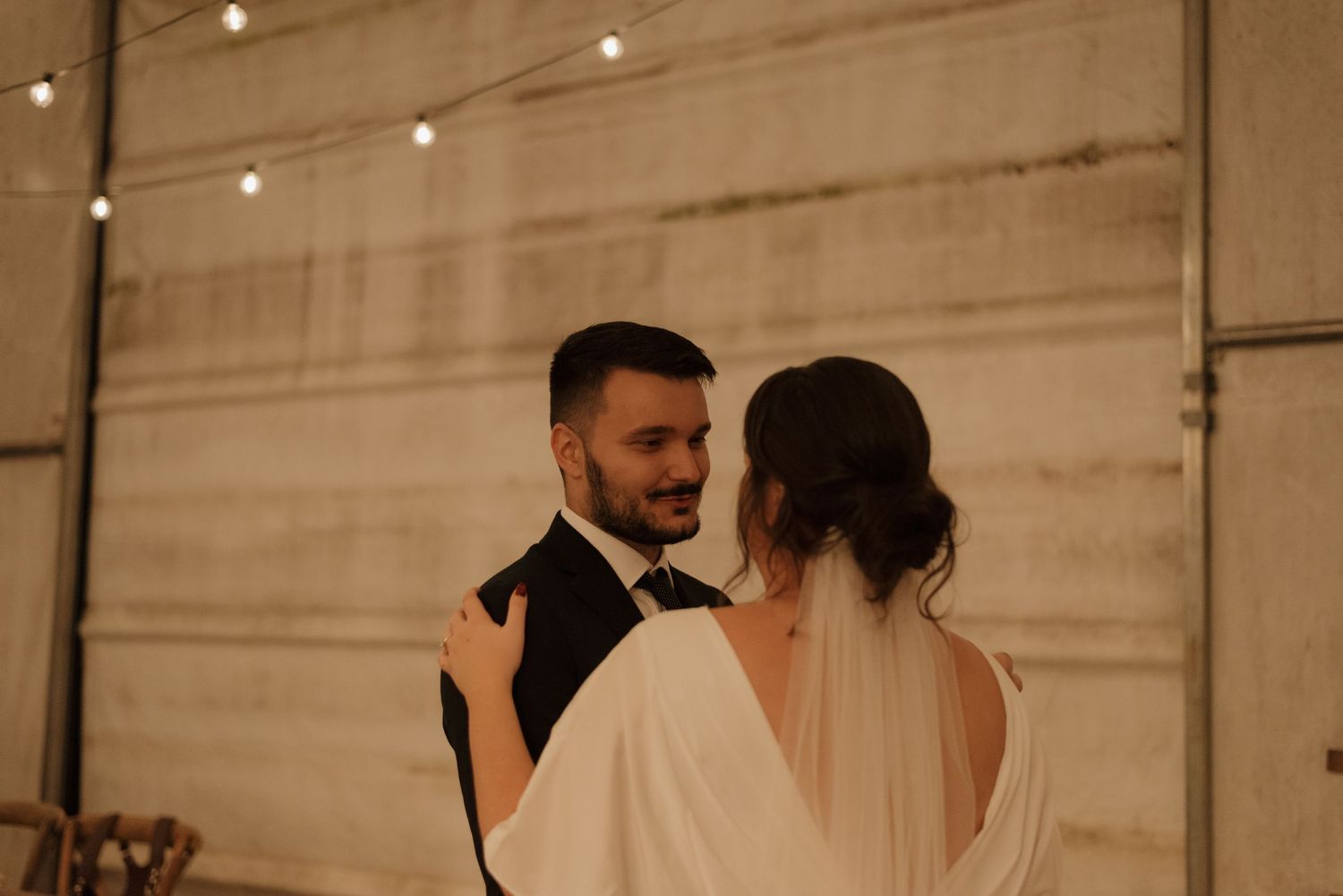 Wedding couple embraces in warm lighting with string lights and rustic backdrop.
