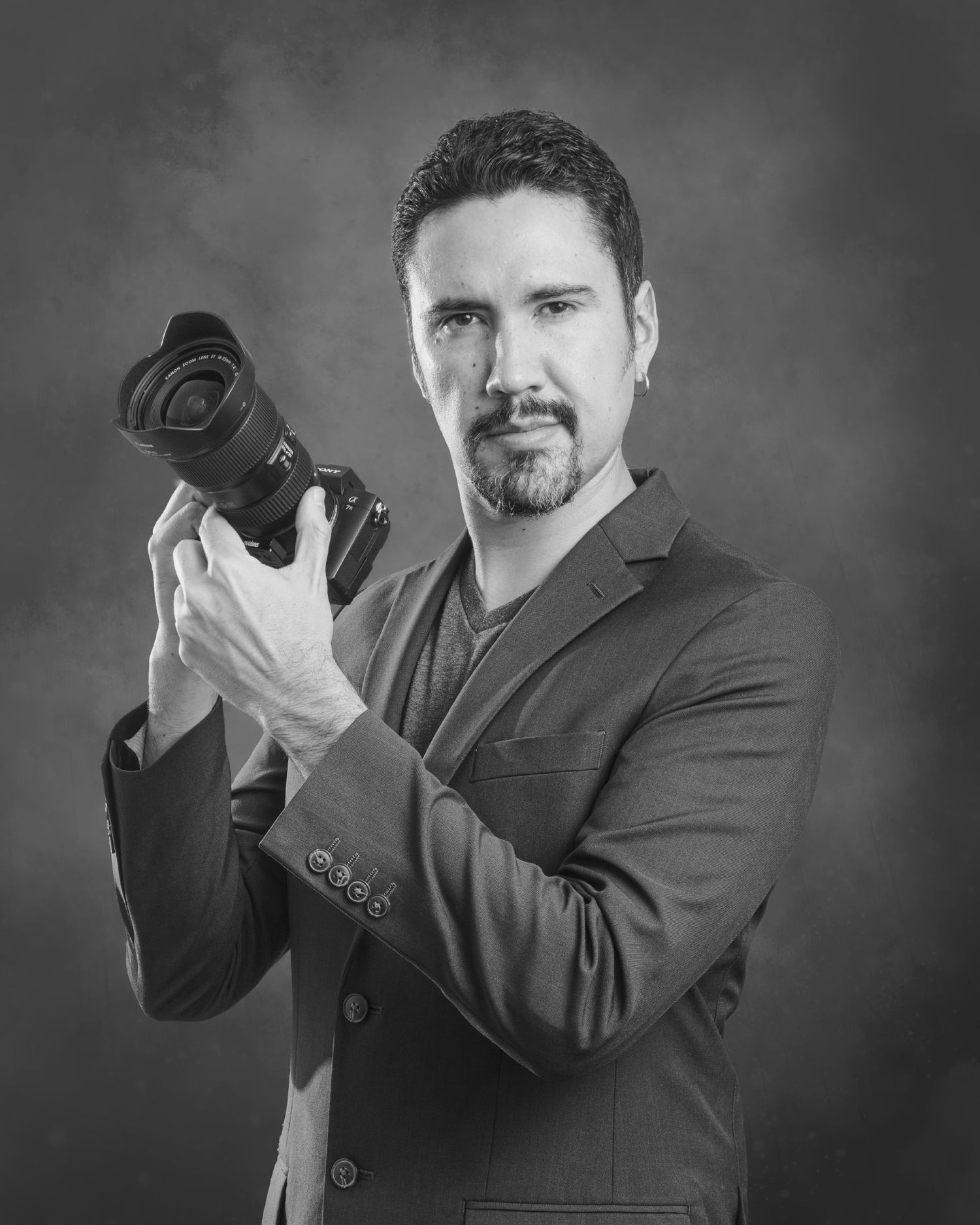 Luis Erantzcani, professional photographer in dark suit holding mirrorless camera against gray background.