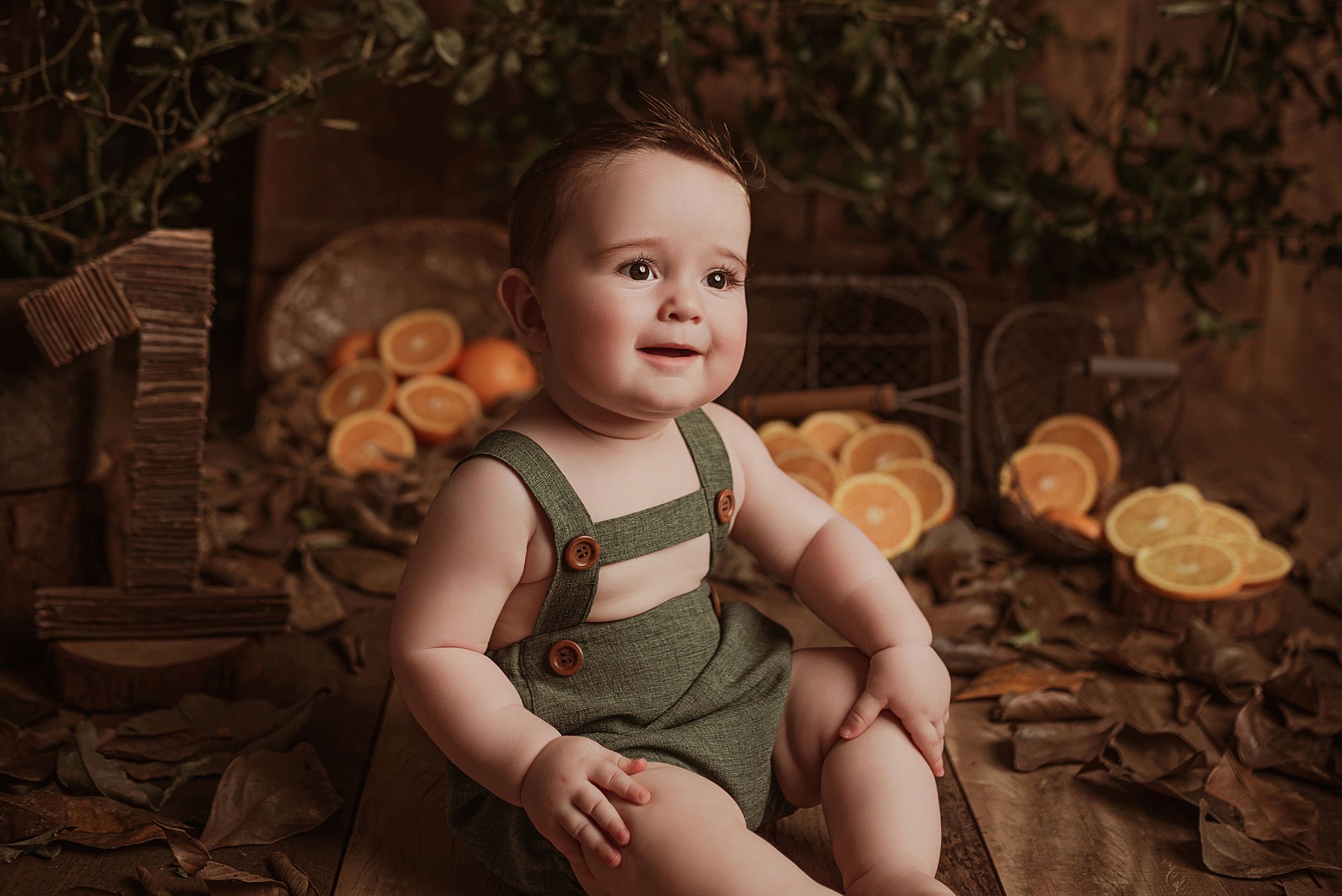 Baby in green overalls sits on wooden floor with glowing orange citrus slices arranged in background.