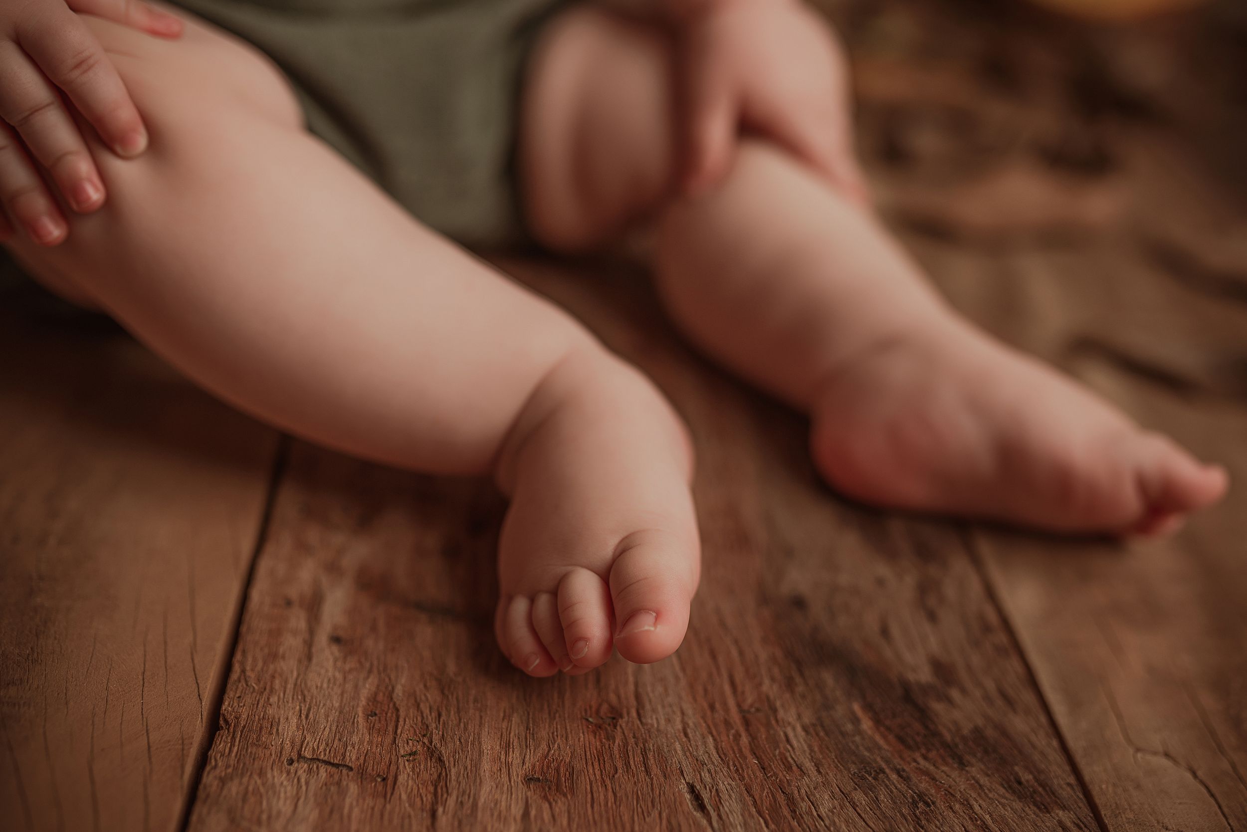 Tiny bare baby feet rest on warm wooden floorboards in soft natural lighting.