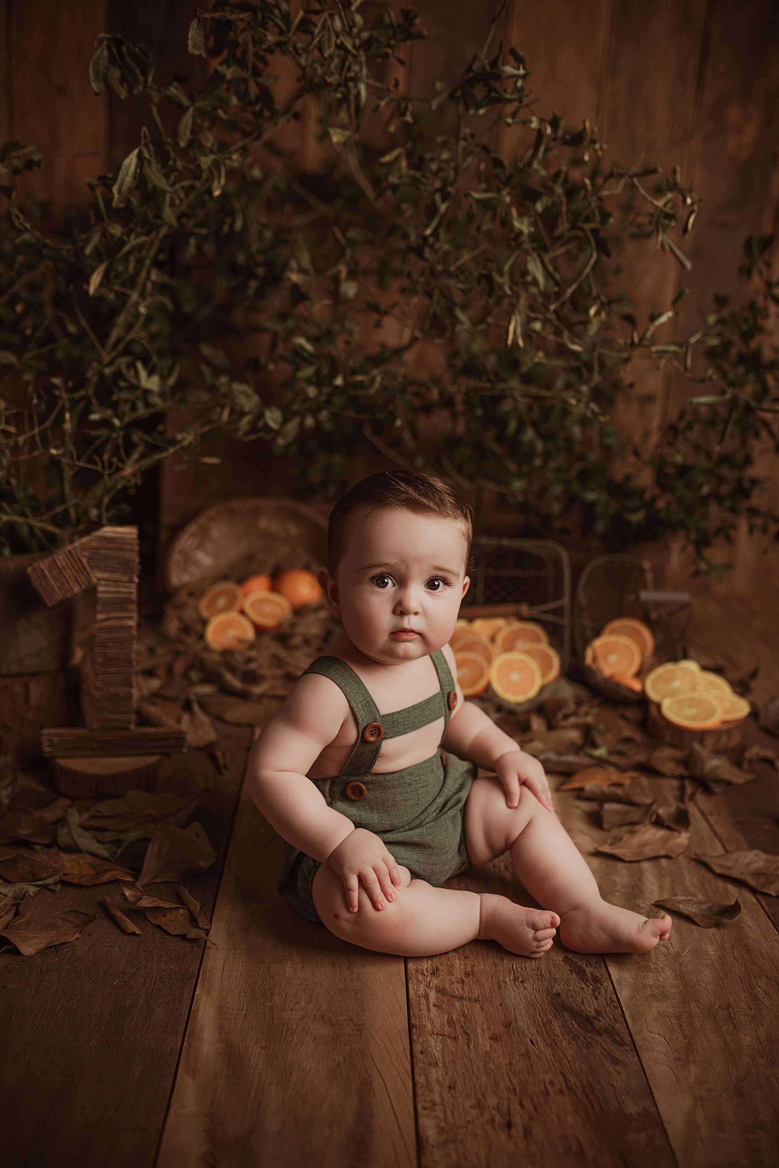 Baby in green overalls sits on rustic wooden floor with citrus fruits and greenery in warm brown background setting.