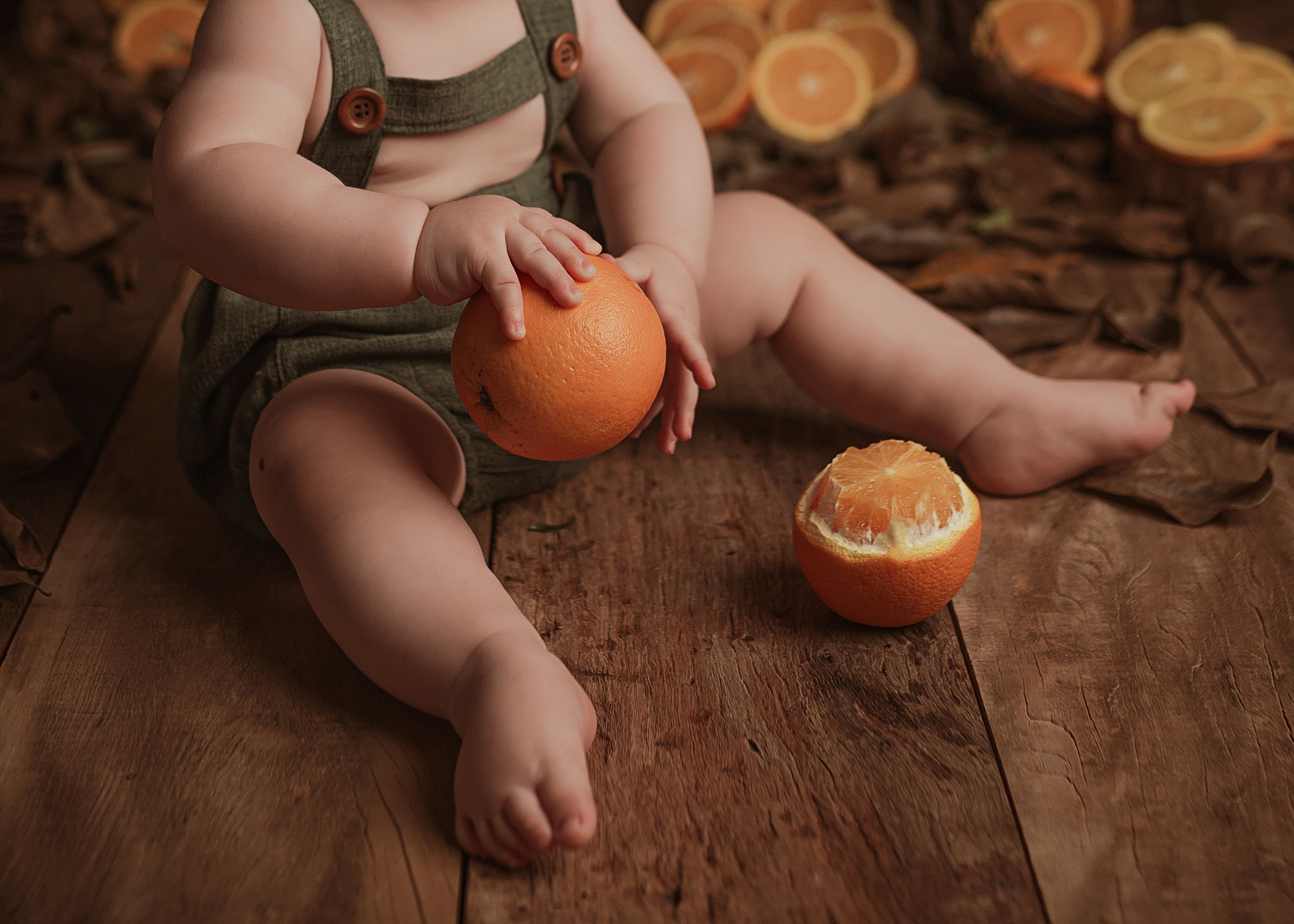 Baby in dark green romper sits on wooden floor playing with fresh oranges at their feet.