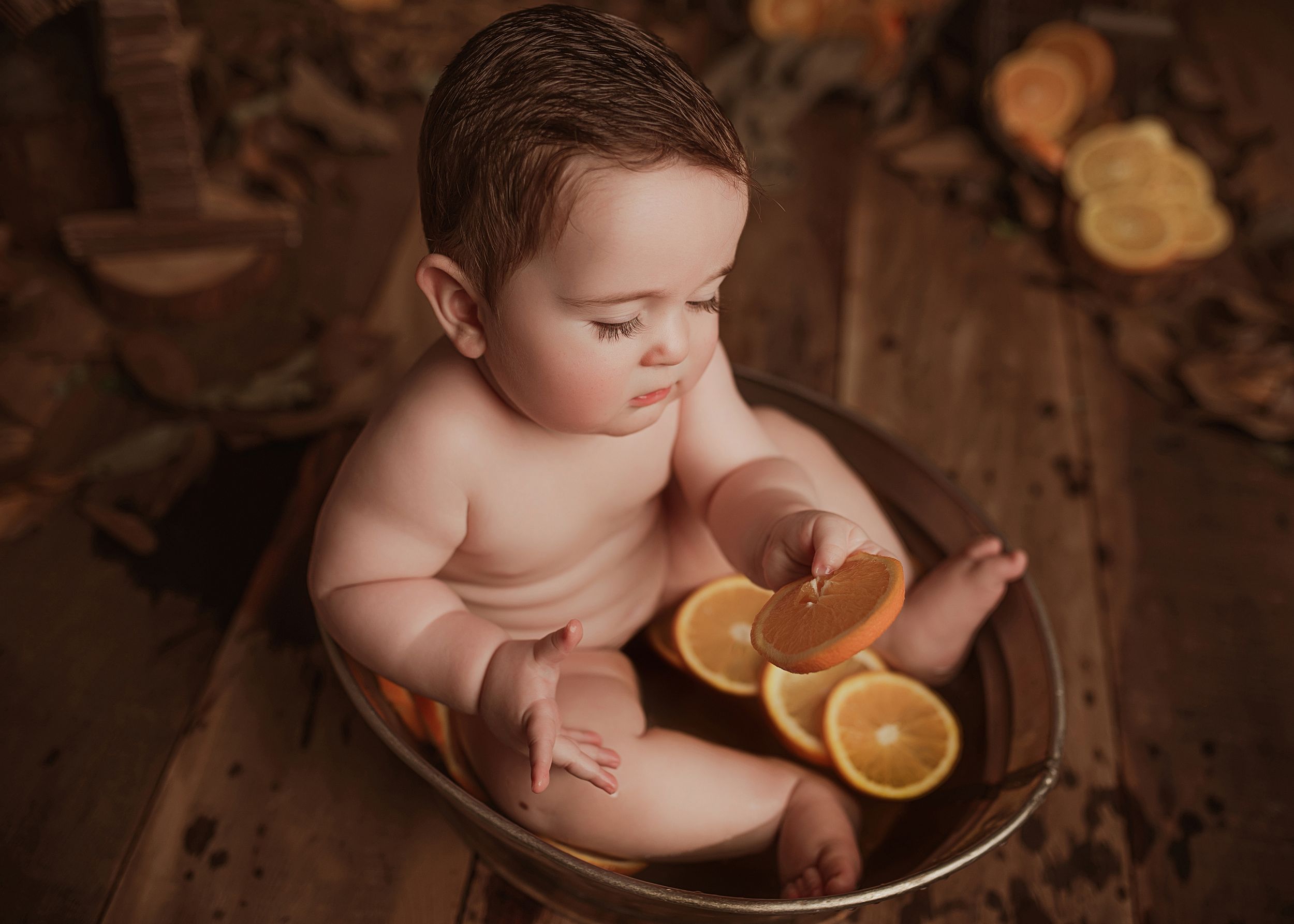 A baby sits in a wooden bowl with orange citrus slices on a dark wooden backdrop for a rustic studio portrait.