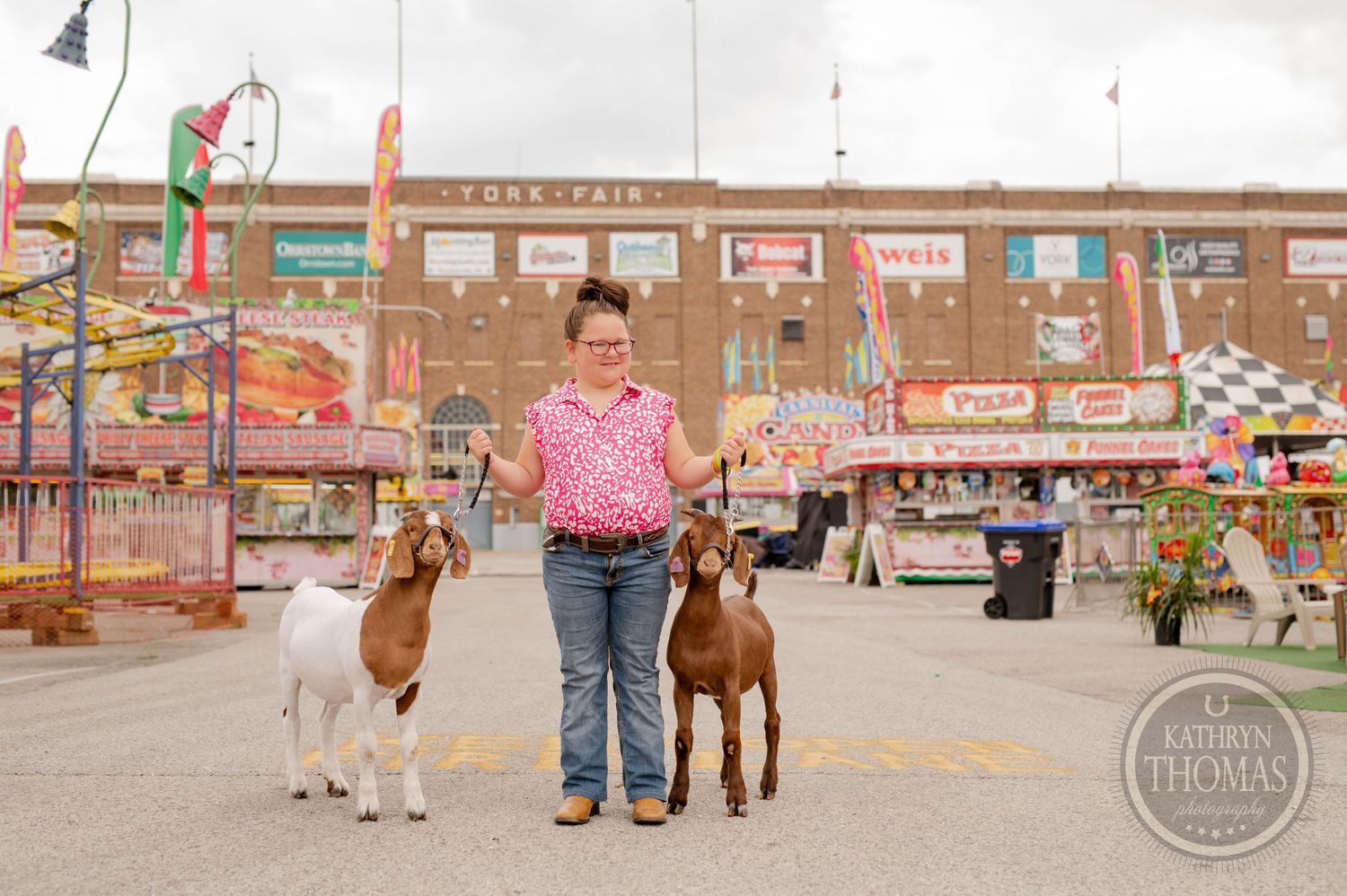 Two goats on leashes stand at a county fair with carnival rides and attractions visible.