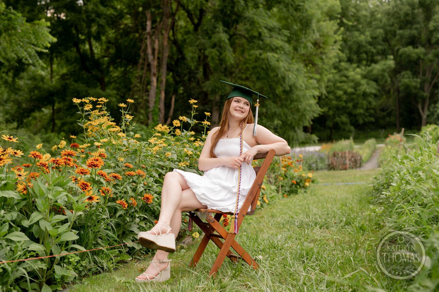 Graduate seated in wooden chair poses near vibrant orange and yellow garden flowers.